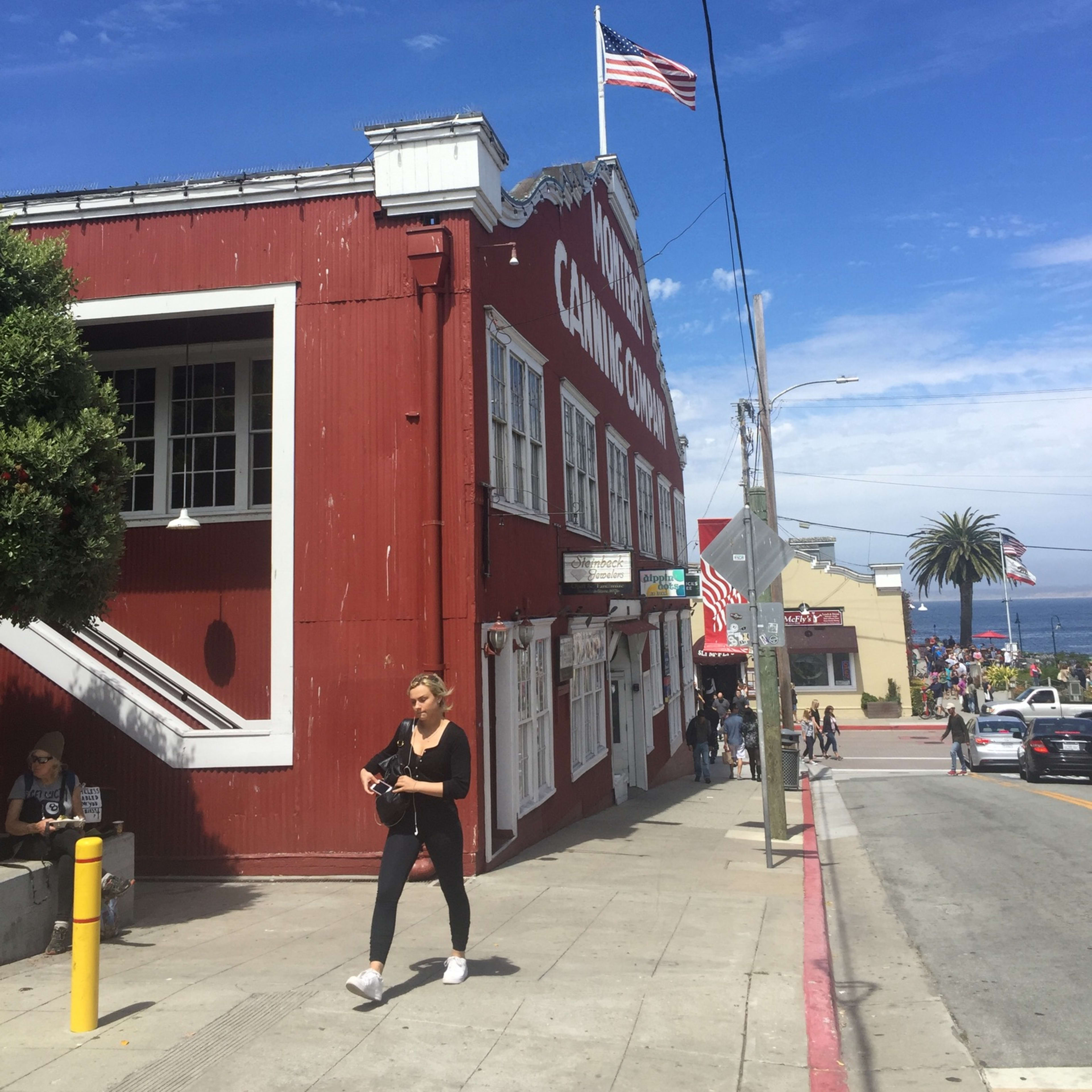 A woman walks past a red building marked "Monterey Canning Company" with an American flag flying above, while the ocean is visible in the background.