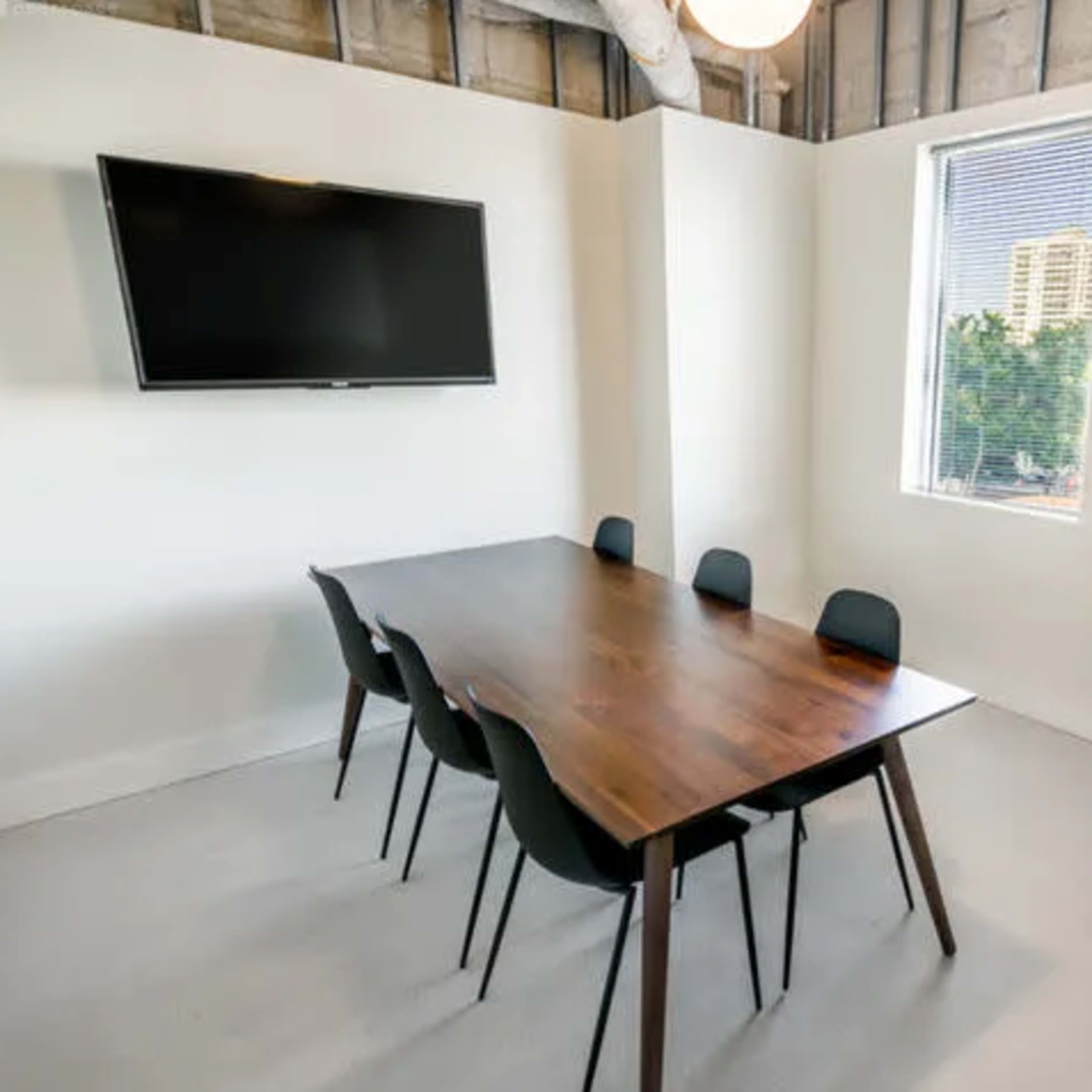 A conference room features a wooden table surrounded by six black chairs, with a wall-mounted television and a window displaying city views.