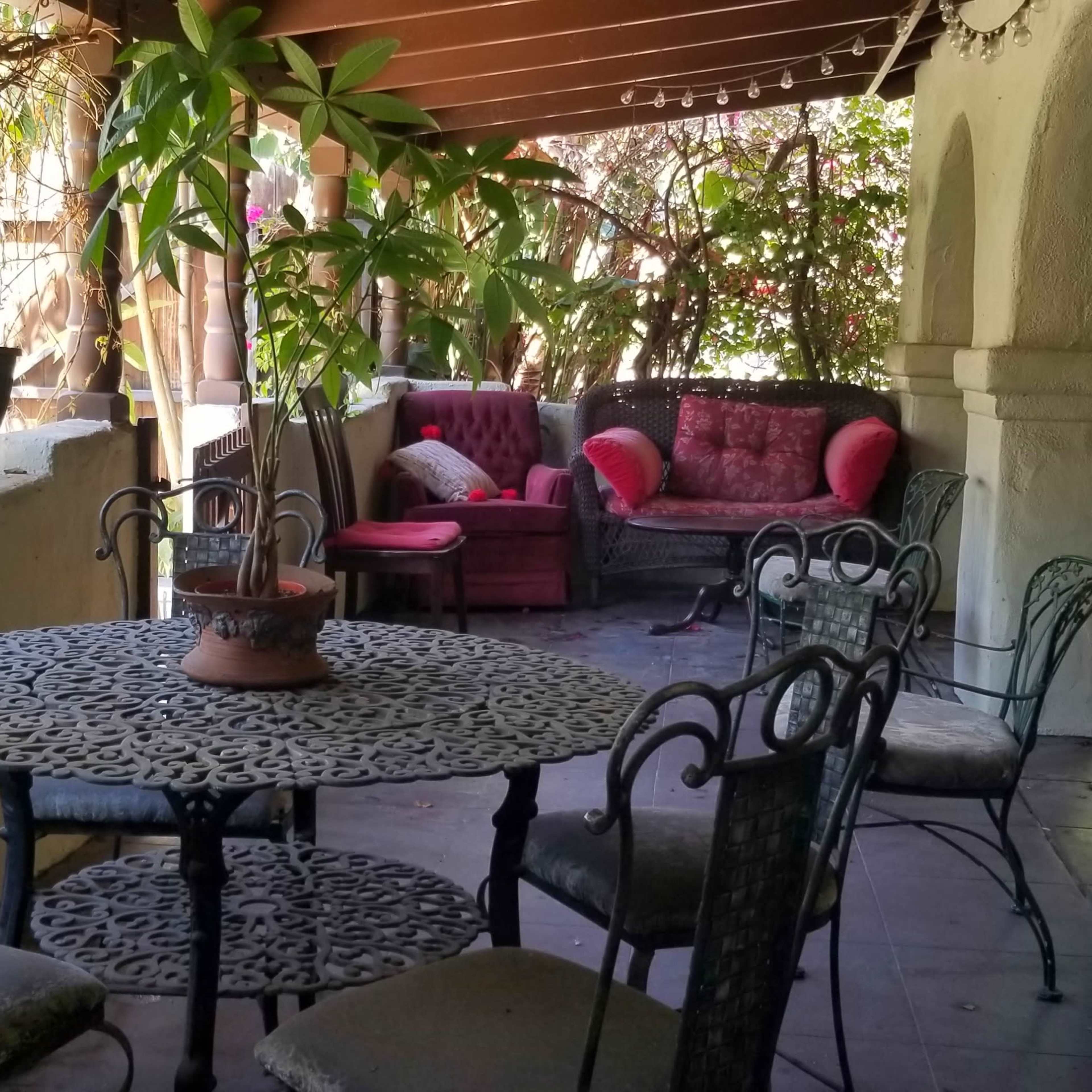A covered outdoor seating area features a wrought-iron table surrounded by six chairs, with plants and comfortable couches in the background.