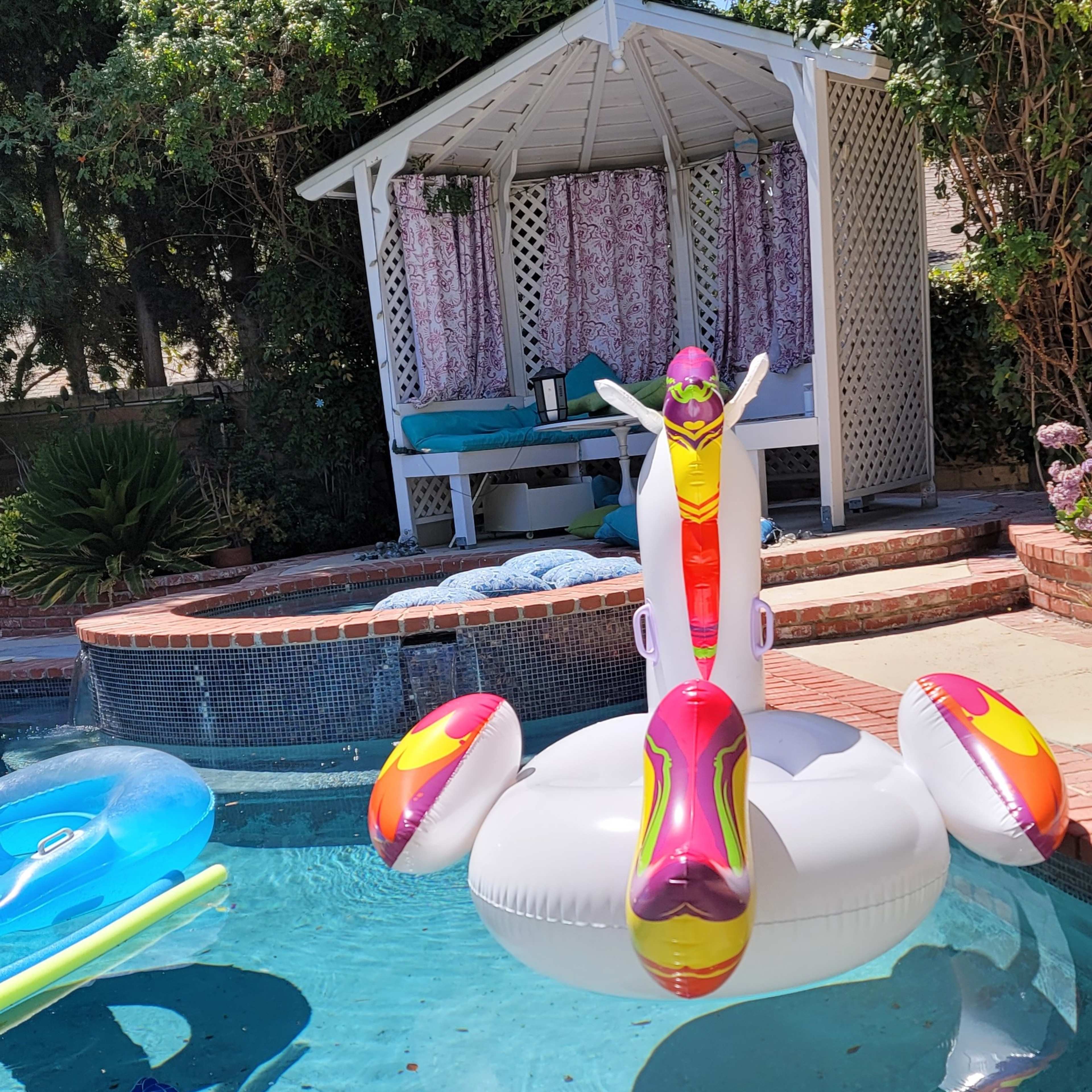 A large inflatable flamingo and a smaller inflatable float are positioned in a swimming pool near a shaded gazebo.