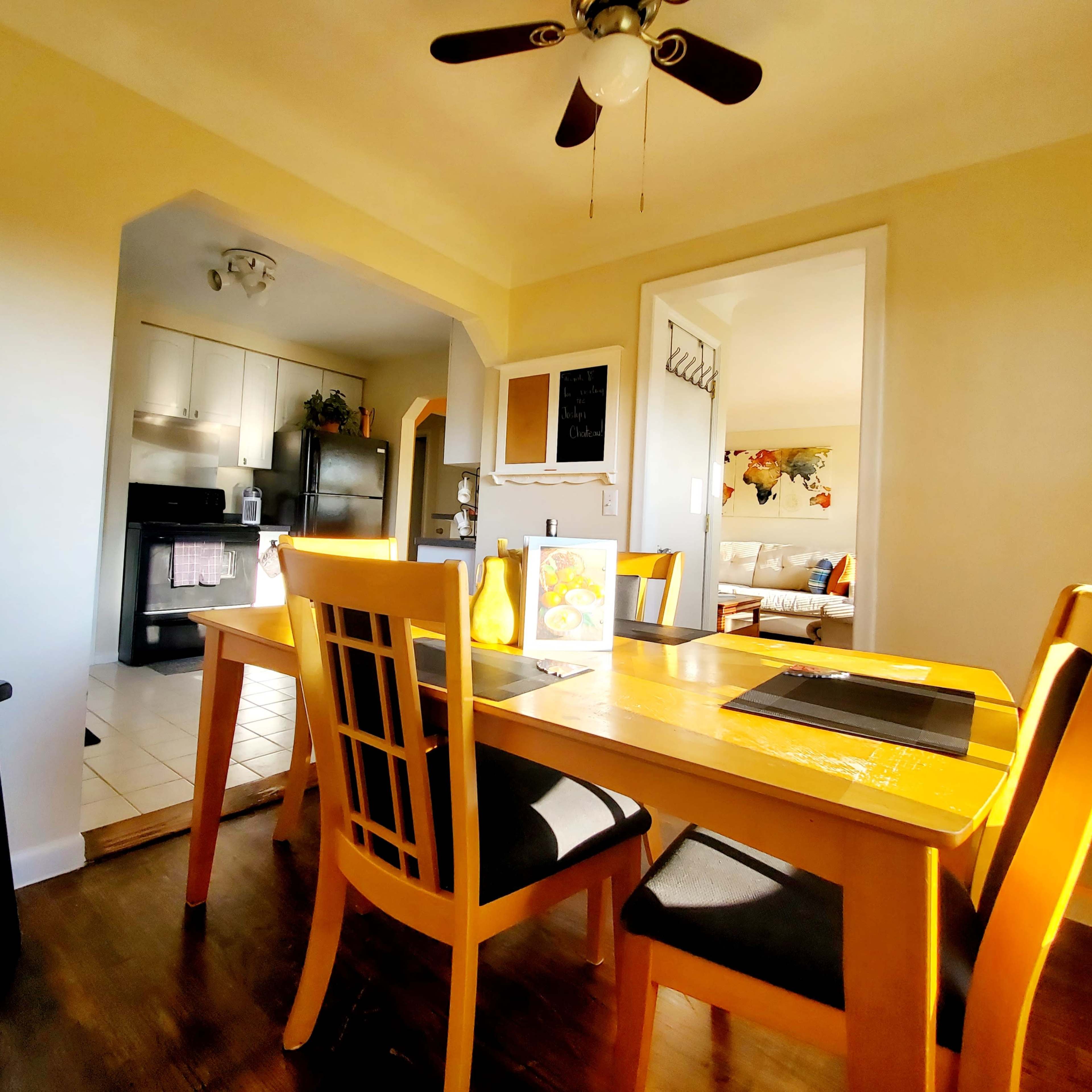 A dining area features a wooden table surrounded by chairs, with a doorway leading to a kitchen and another room in the background.