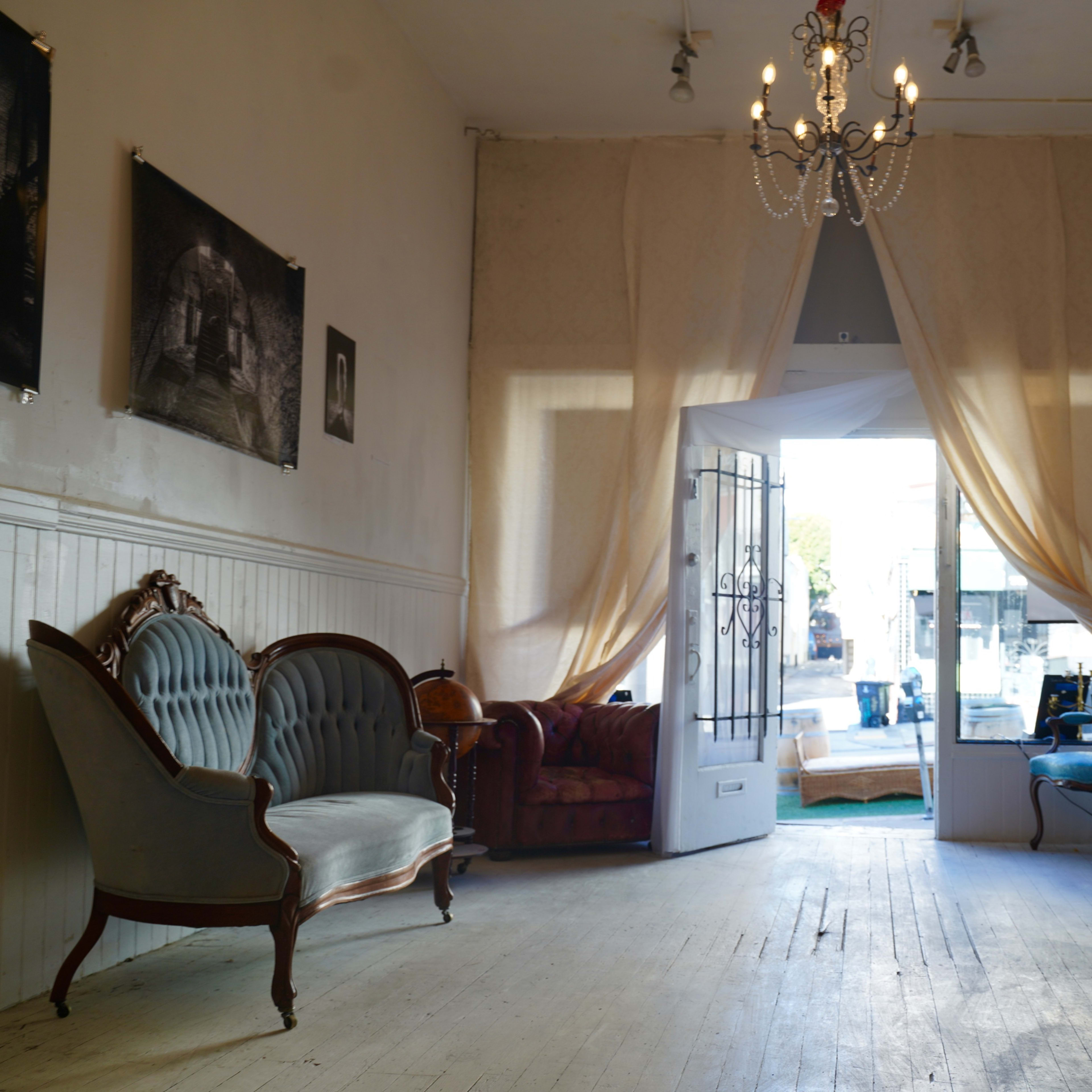 A lightly furnished room with two vintage sofas, a piano, and an open door leading outside, adorned with soft drapes and framed photographs on the walls.
