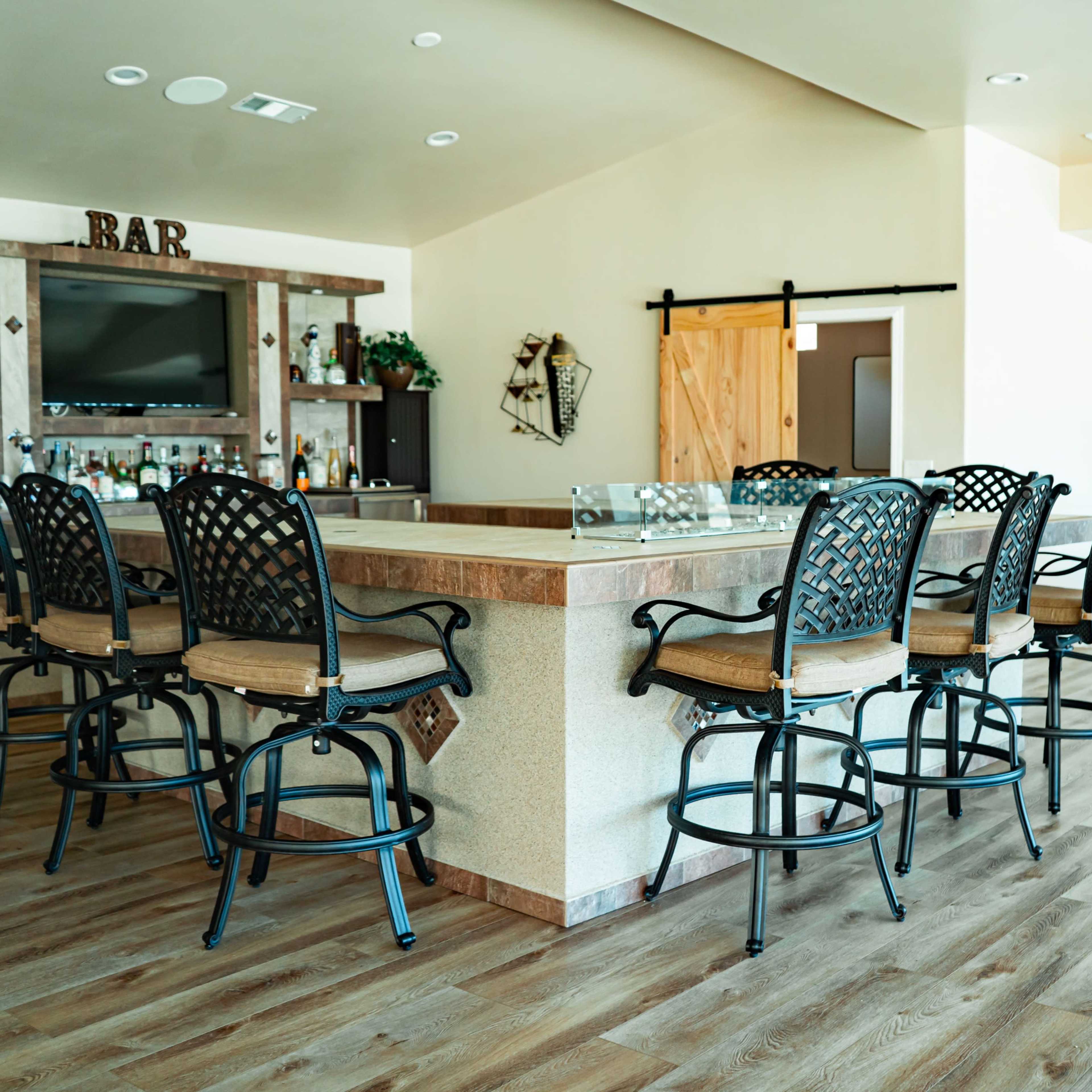 A modern bar area featuring high stools along a countertop, with a television and shelves stocked with beverages in the background.