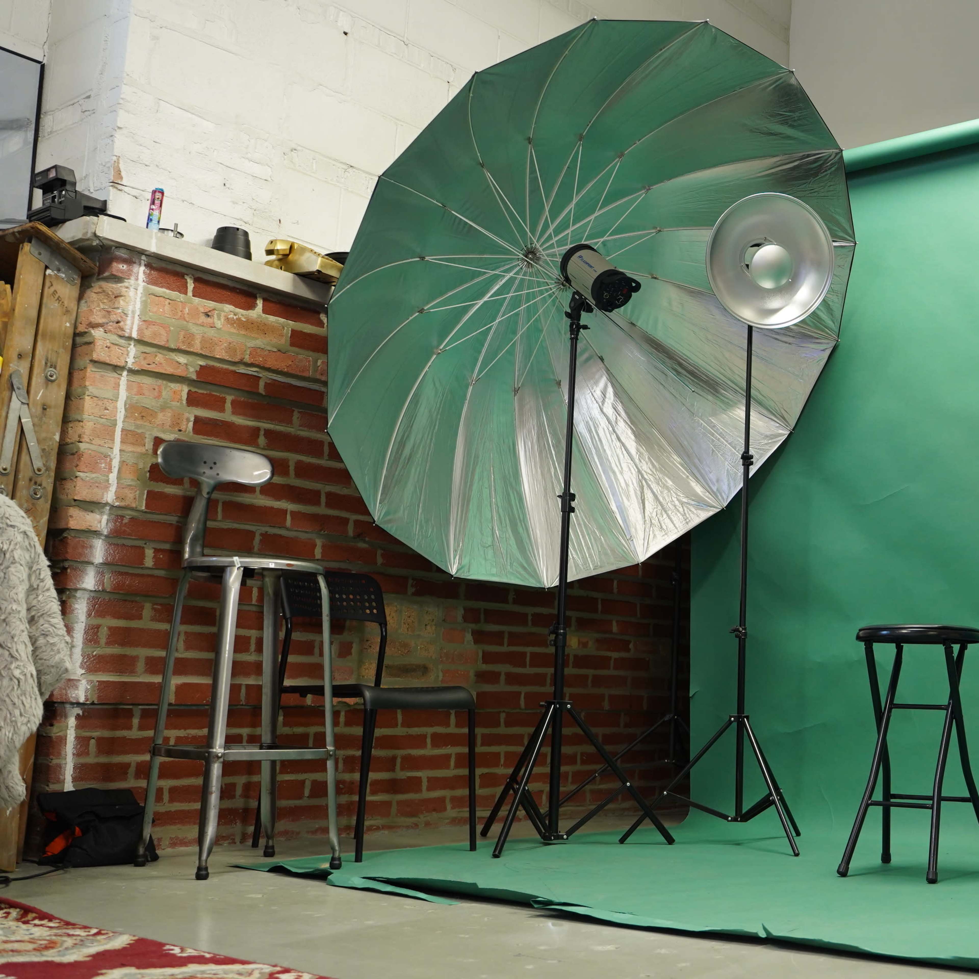 A photography studio equipped with a large umbrella light, a green backdrop, a stool, and a mirror.