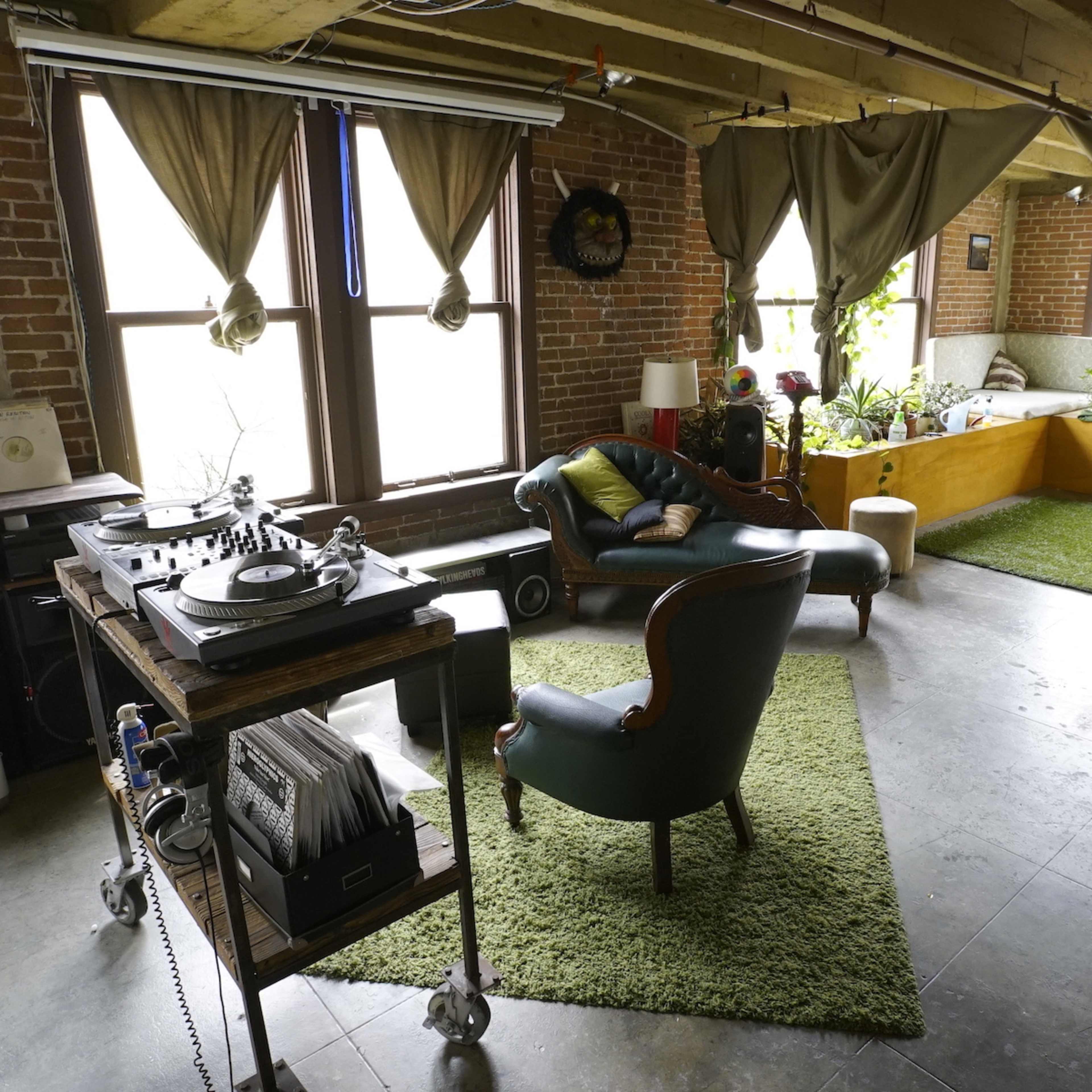 The spacious room features large windows with draped curtains, a vinyl record player on a cart, and a mix of seating arrangements along with plants and a soft green rug.