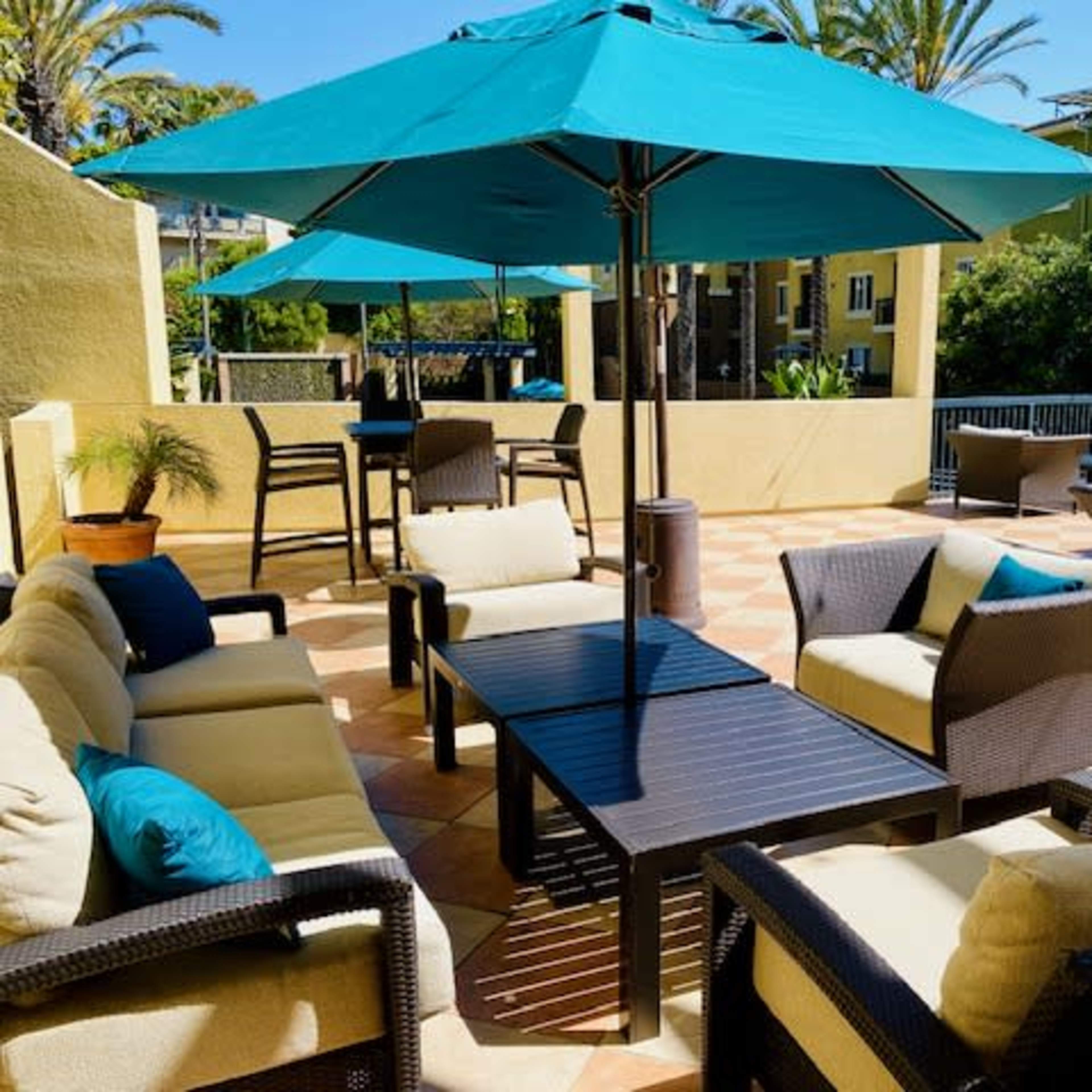An outdoor lounge area with turquoise umbrellas, wicker seating, and a tiled floor surrounded by palm trees and buildings.