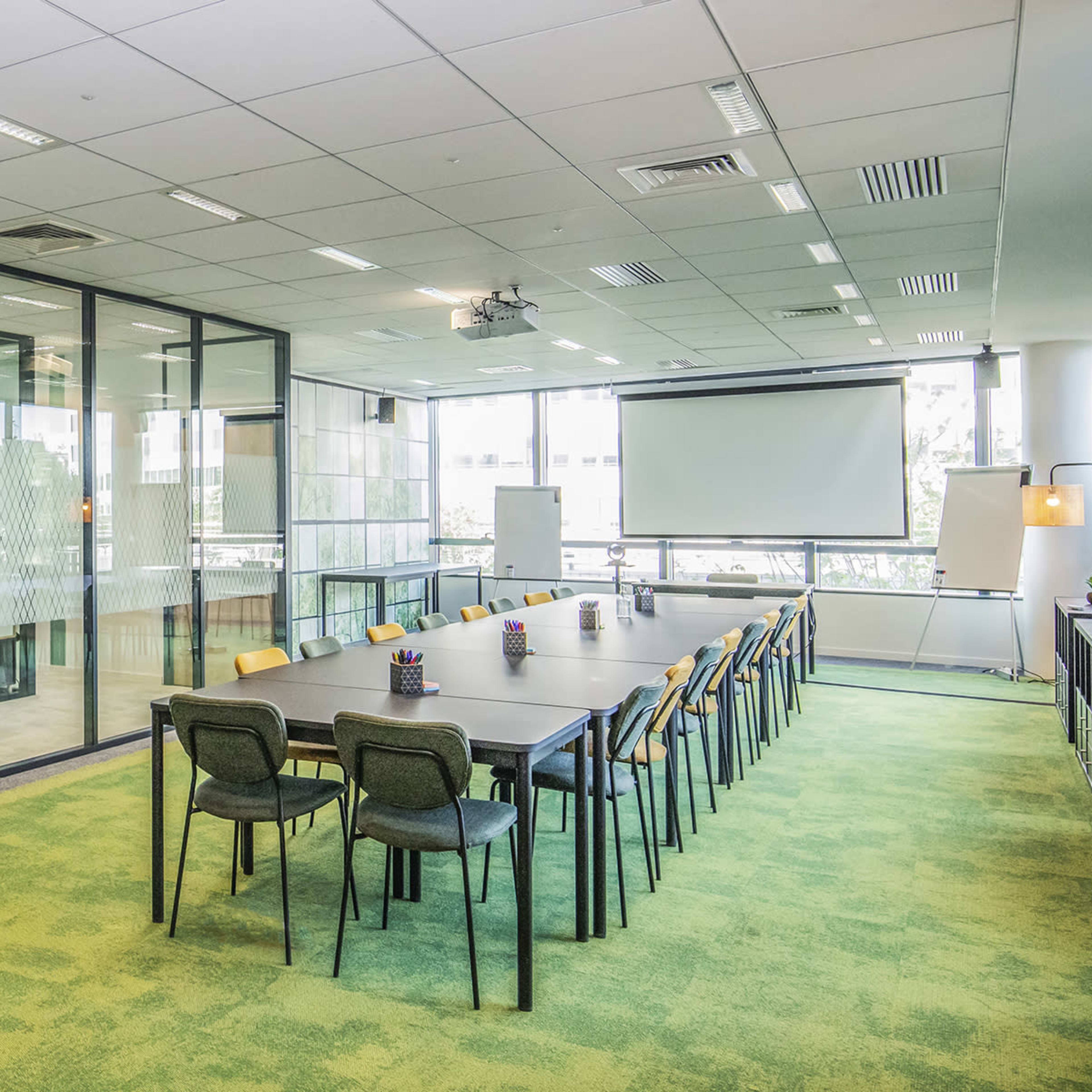 A modern conference room with a long table surrounded by chairs, large windows, and a projector screen at one end.