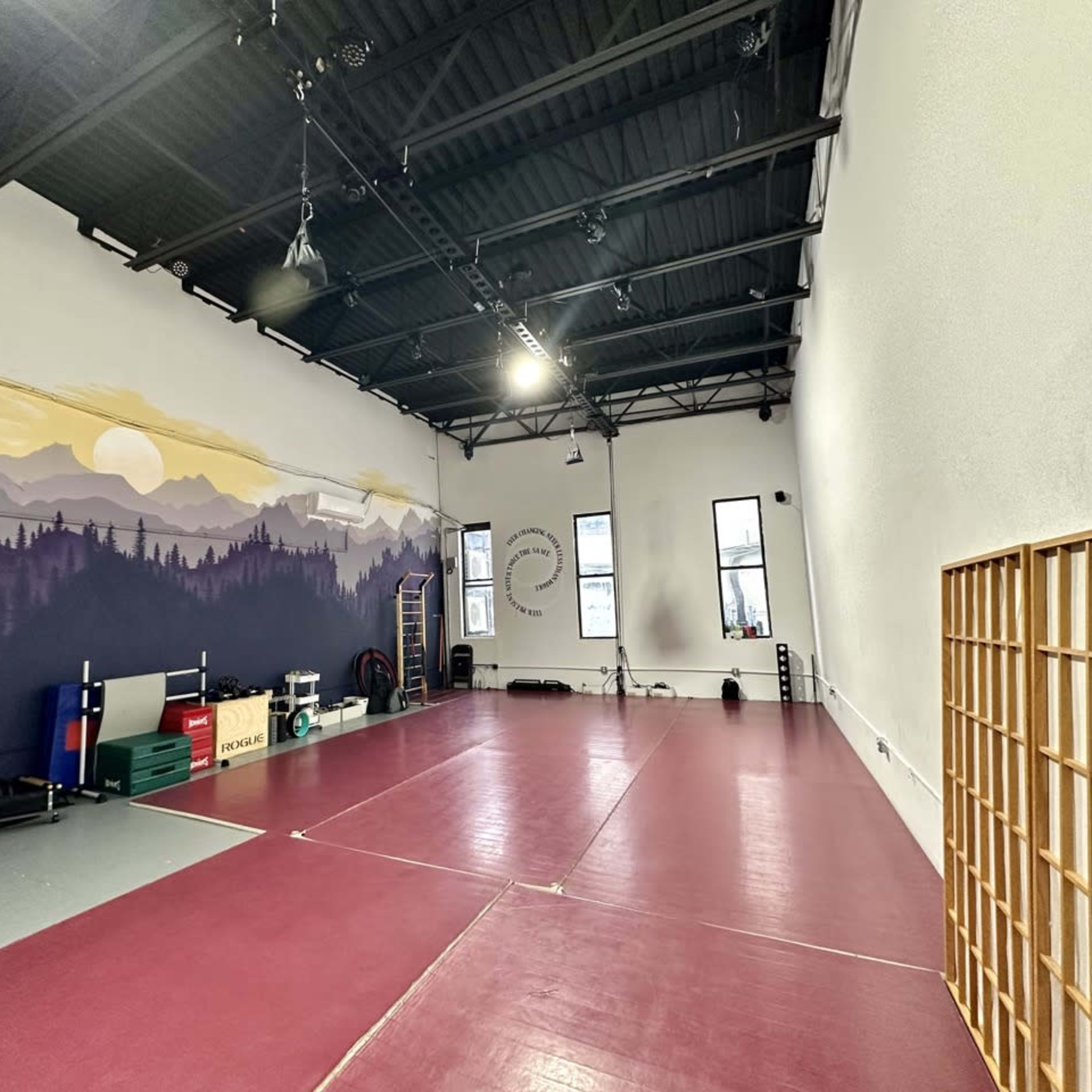 A spacious indoor martial arts studio with a red rubber mat, large windows, and a mural of mountains on one wall.