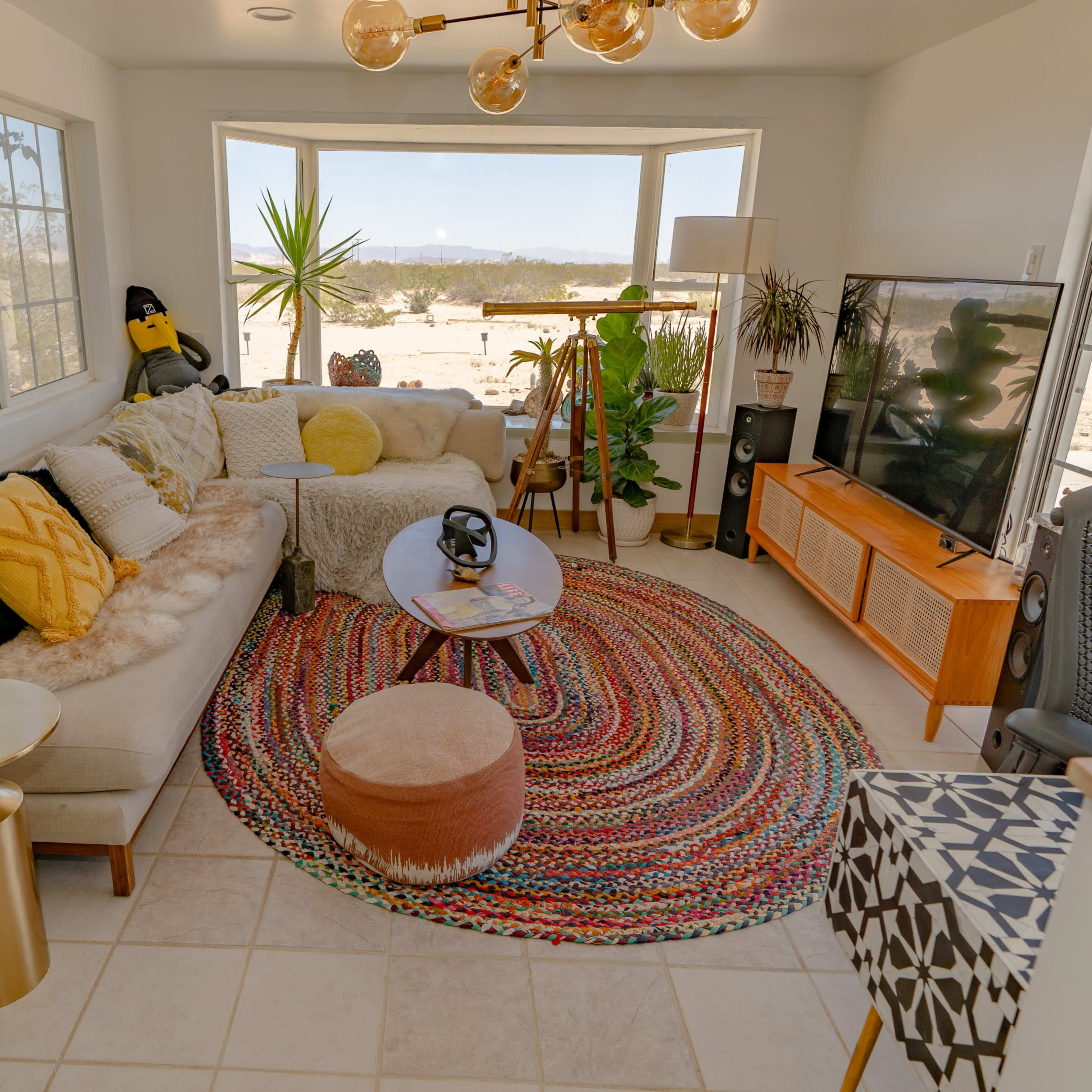 A brightly lit living room features a sectional sofa, a round coffee table, and large windows overlooking a desert landscape, surrounded by various plants and decorative furnishings.