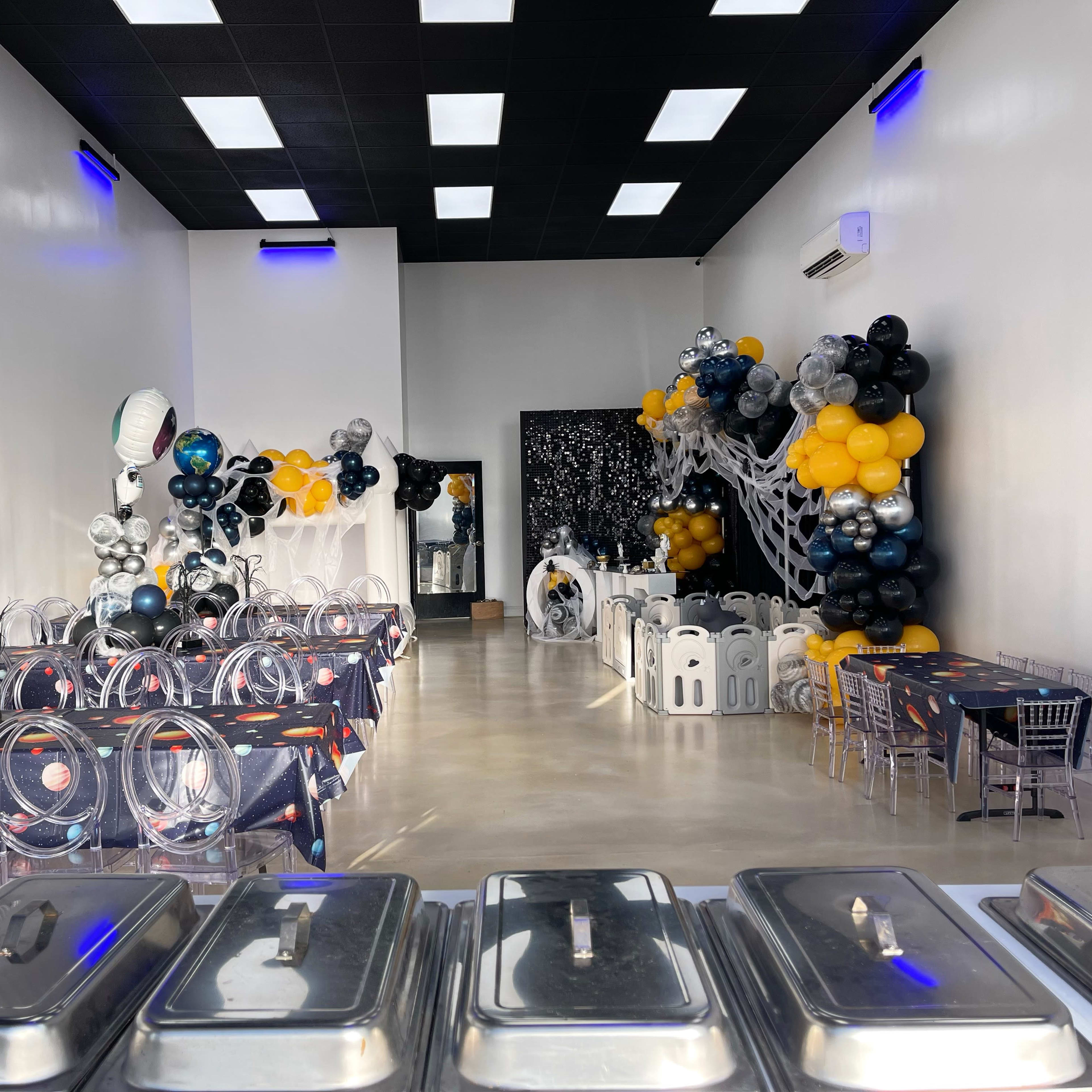 A spacious event hall decorated with space-themed balloons, tables covered with cosmic tablecloths, and a buffet setup in the foreground.