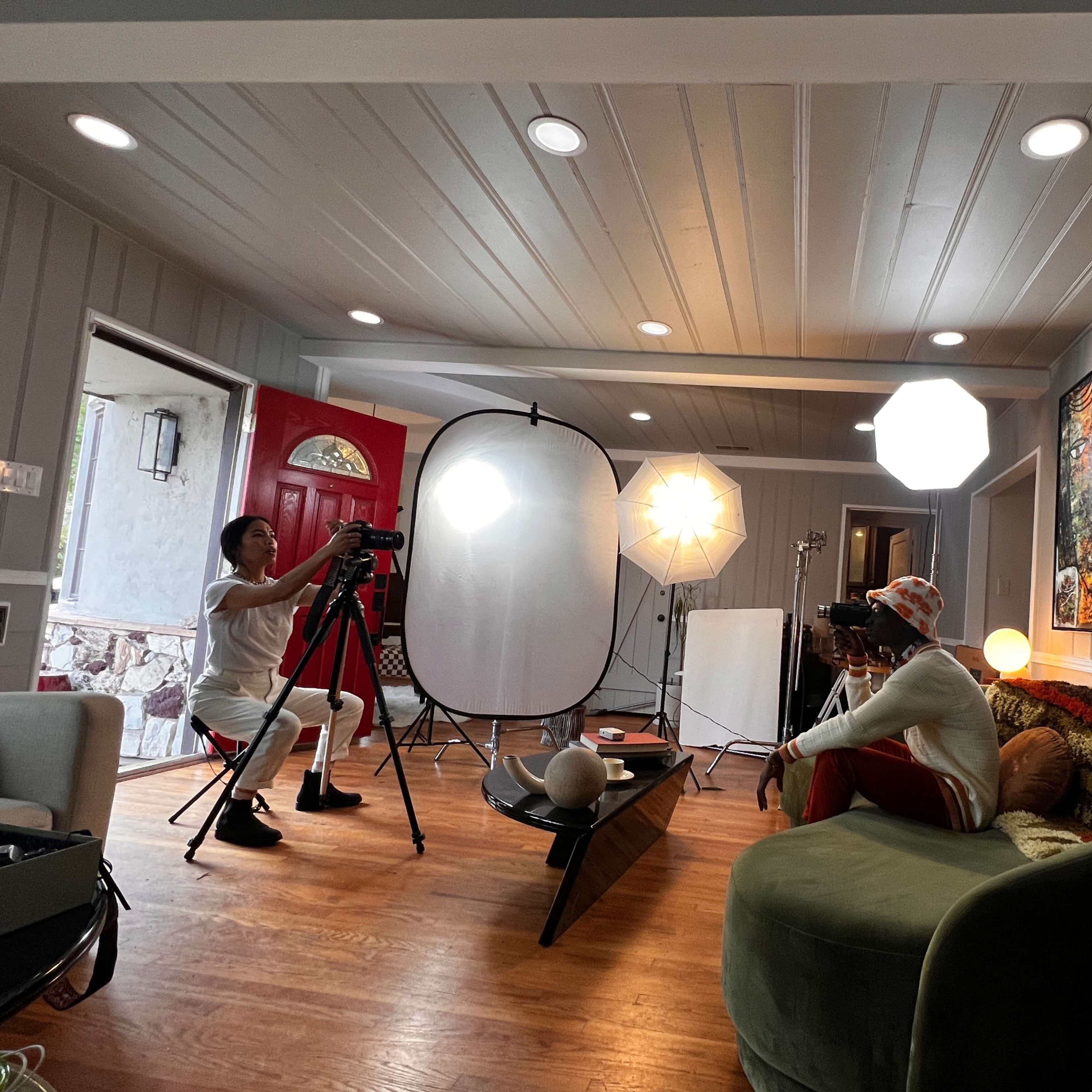 A photographer is capturing images of a model in a well-lit living room featuring a green couch and various lighting equipment.