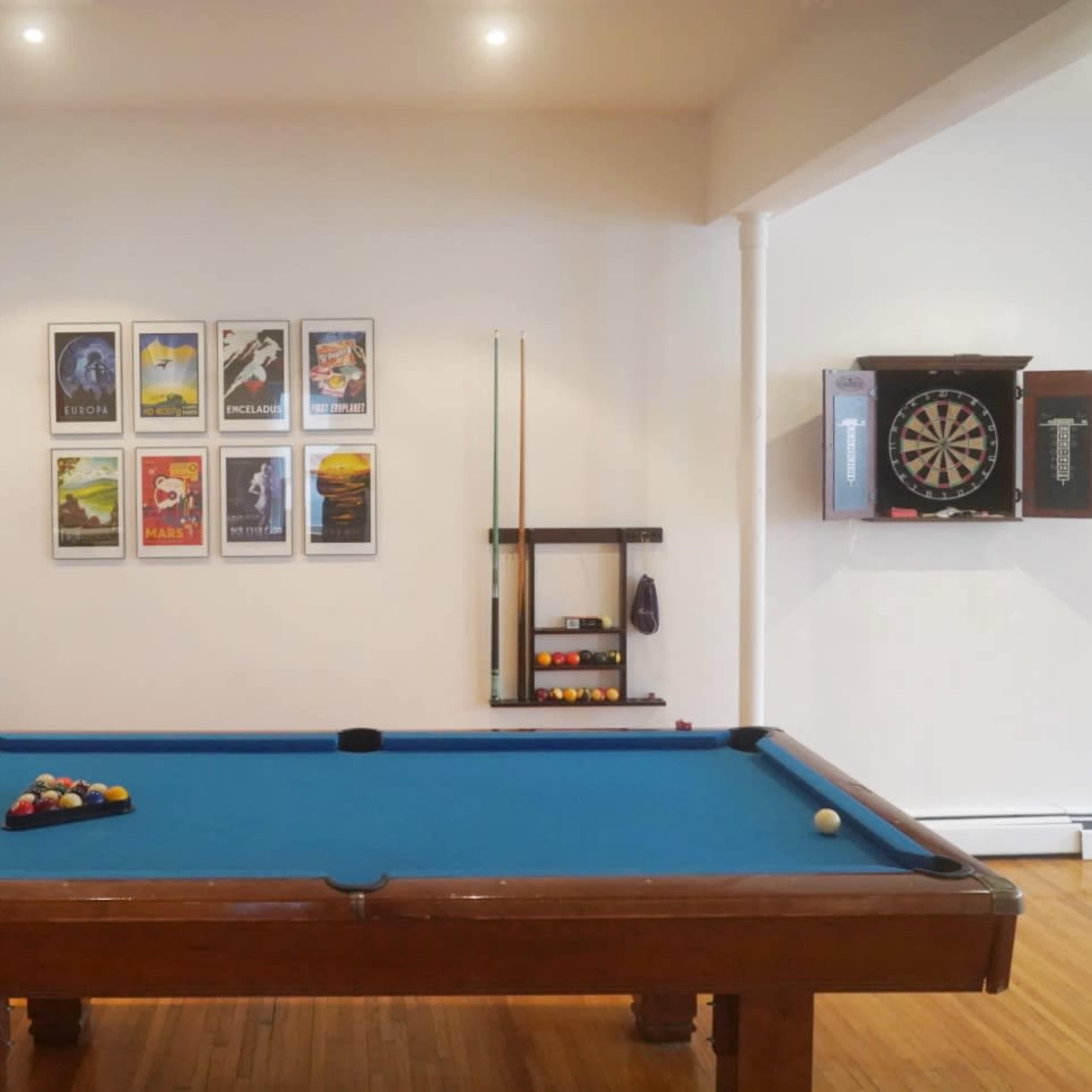 A pool table with a blue felt surface is placed in a room featuring framed posters on the wall and a dartboard on the opposite side.