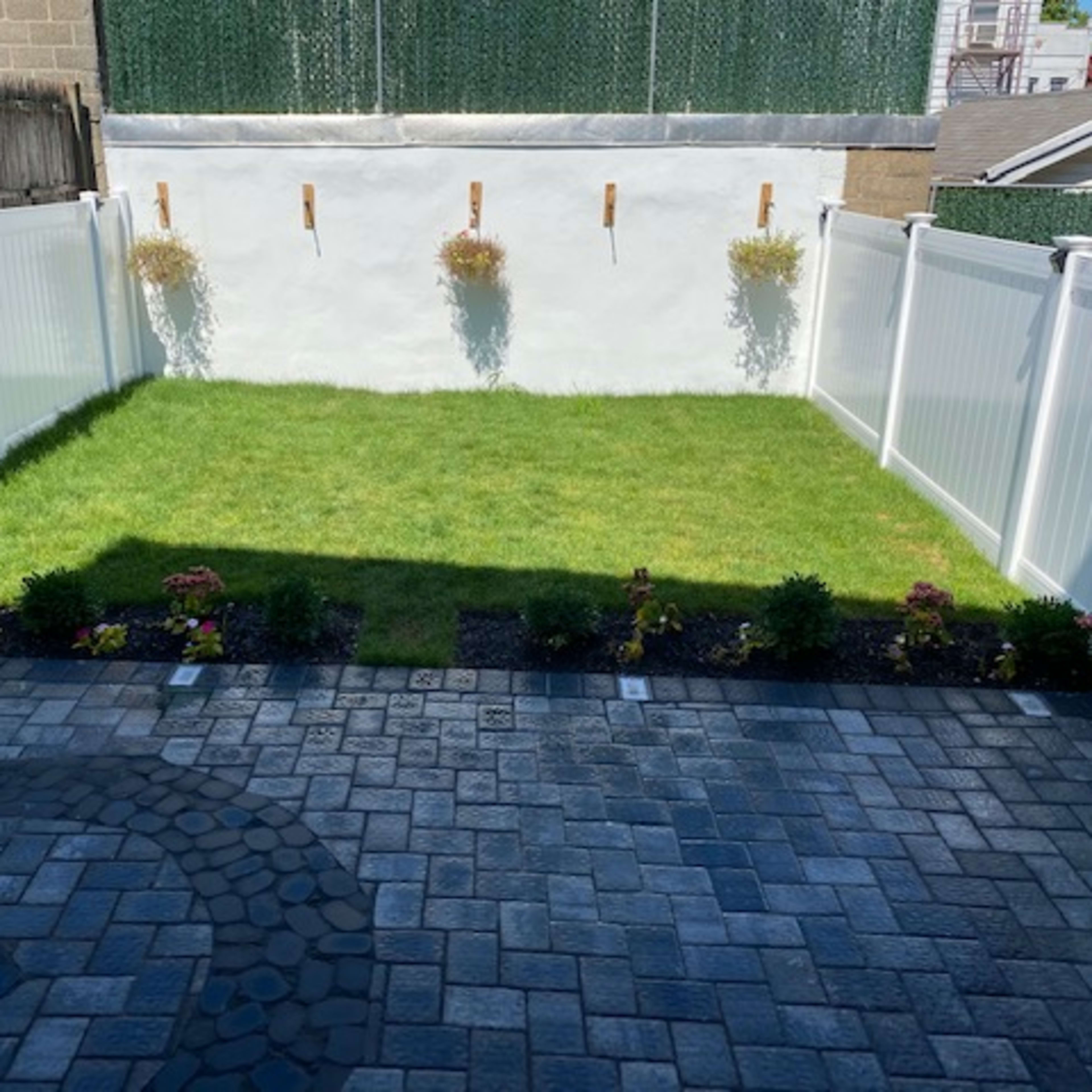 A small, fenced backyard with a grassy area surrounded by a stone patio and flower beds.