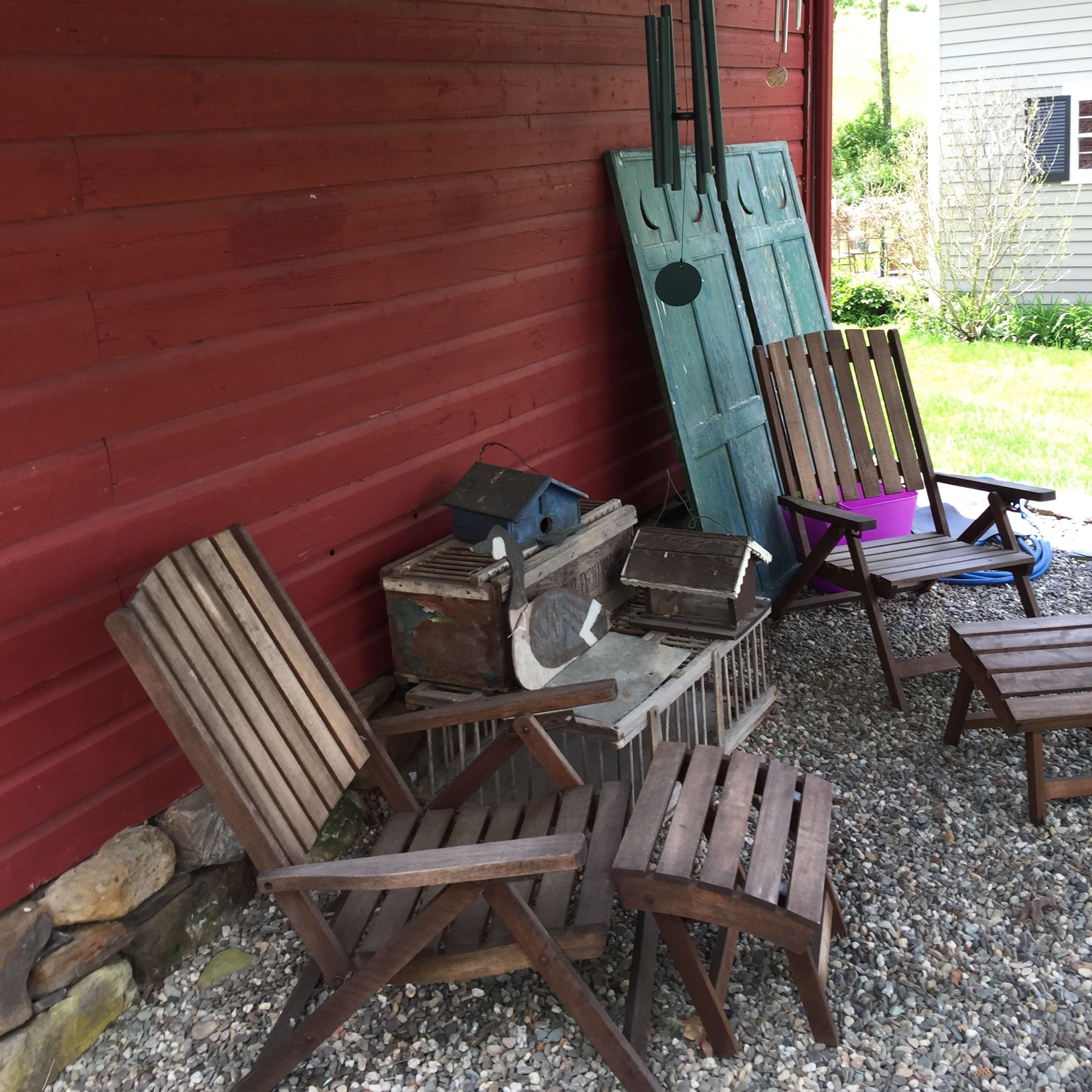 A collection of wooden chairs, a stone table, and various rustic items are arranged under a porch next to a red wall.