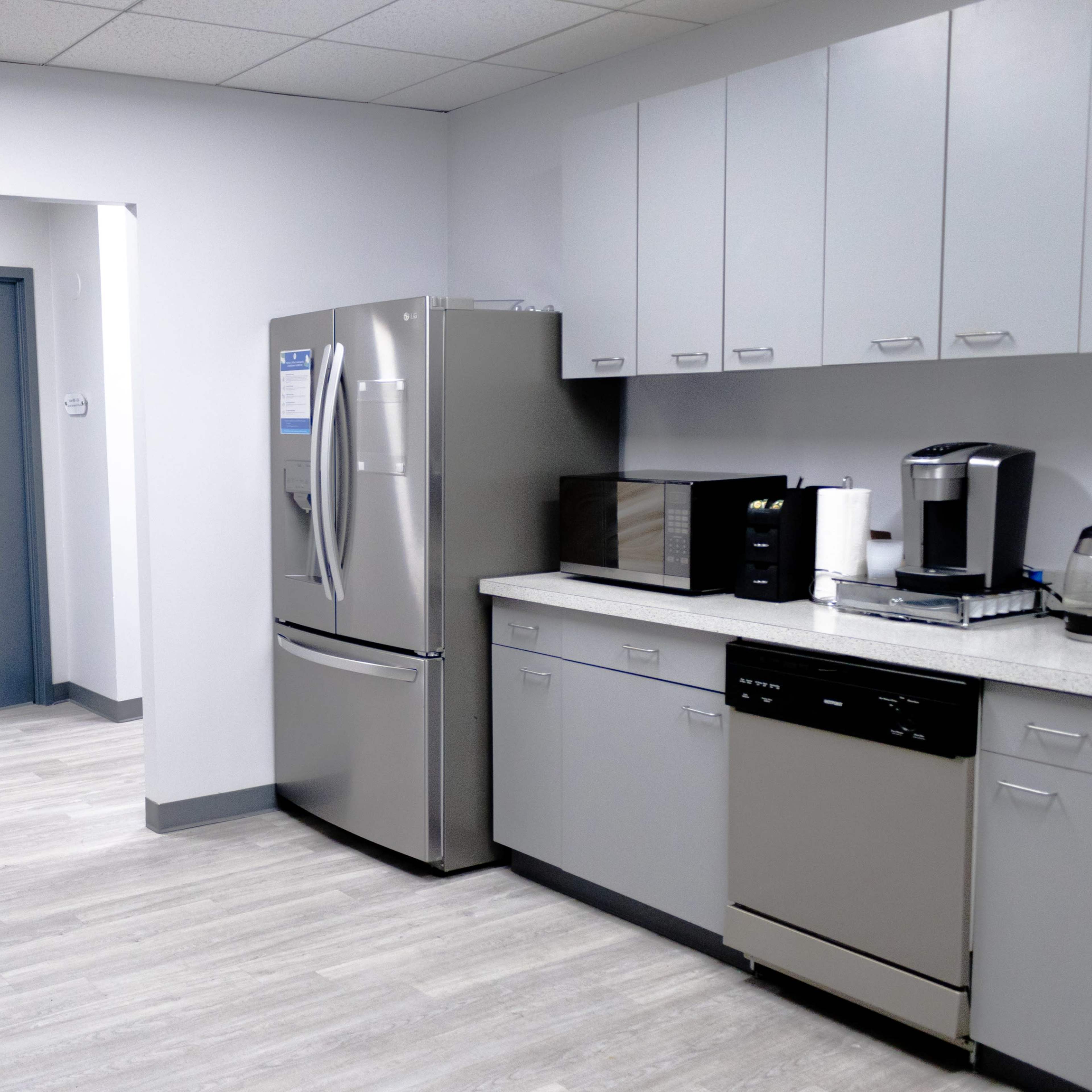 A modern break room with a refrigerator, microwave, coffee maker, and a table with chairs, along with a doorway leading to another room.