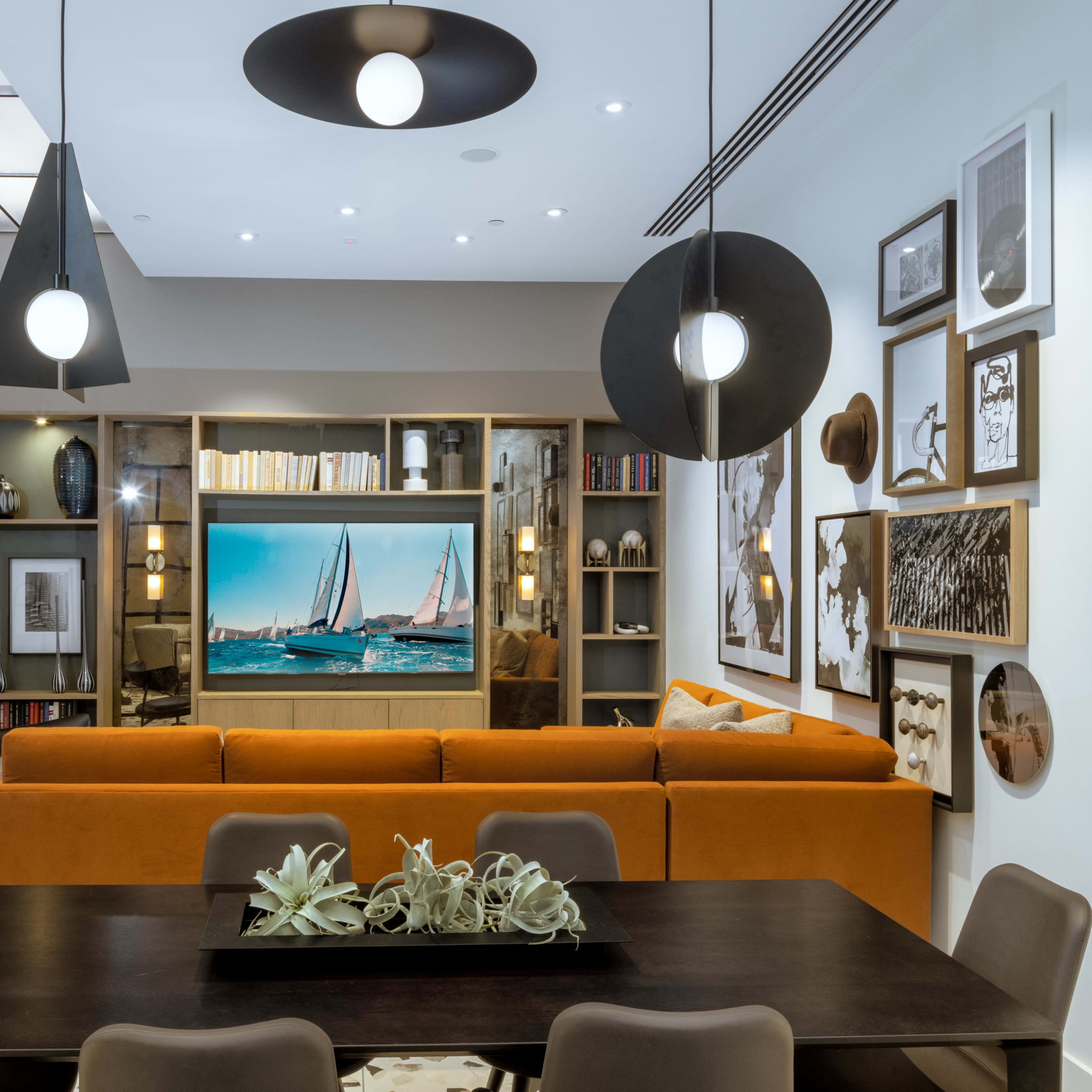 A modern living area with an orange sofa, a dining table, and a large TV unit displaying sailing boats, accented by decorative wall art.