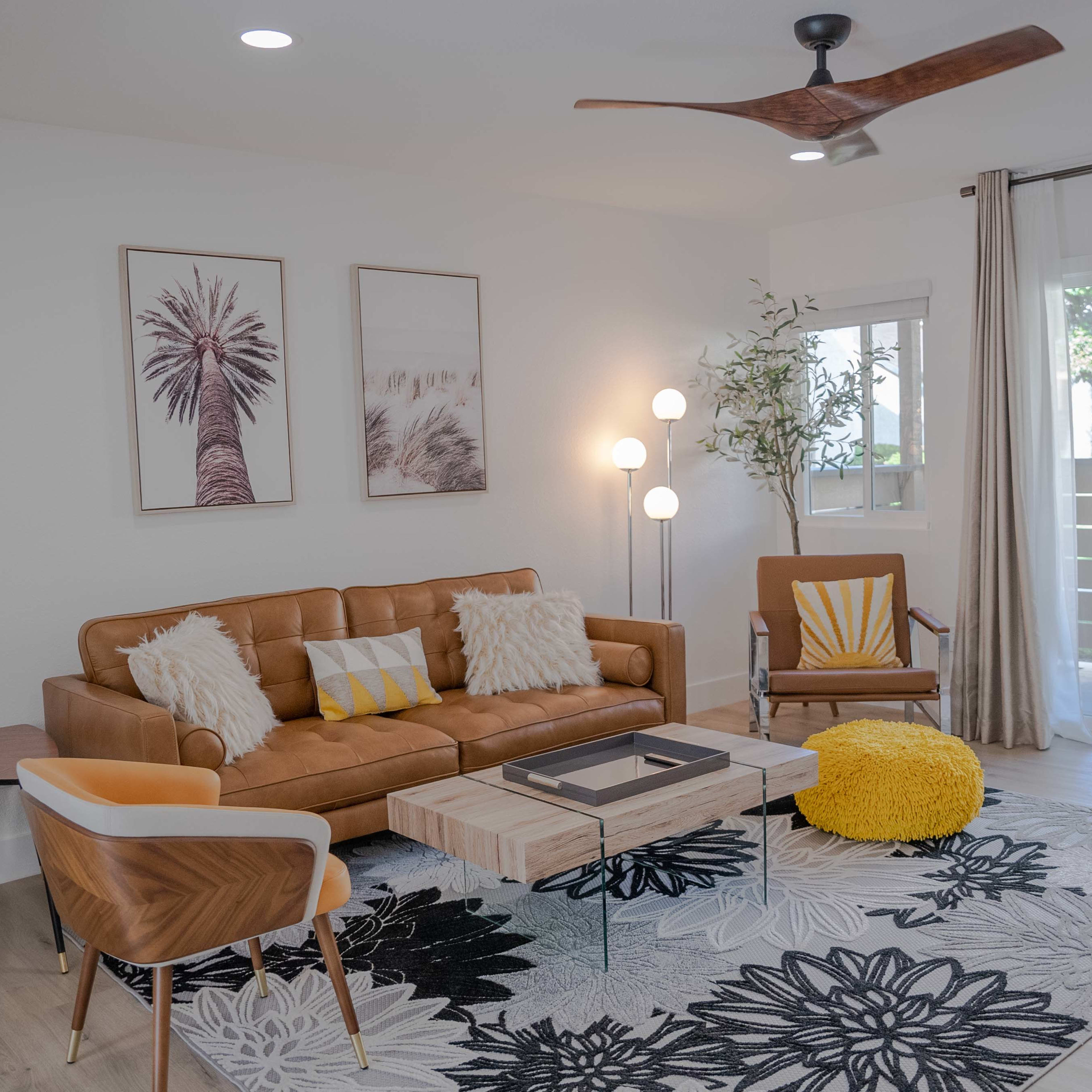 The living room features a brown leather sofa, a wooden coffee table, a patterned rug, and large windows with sliding glass doors.