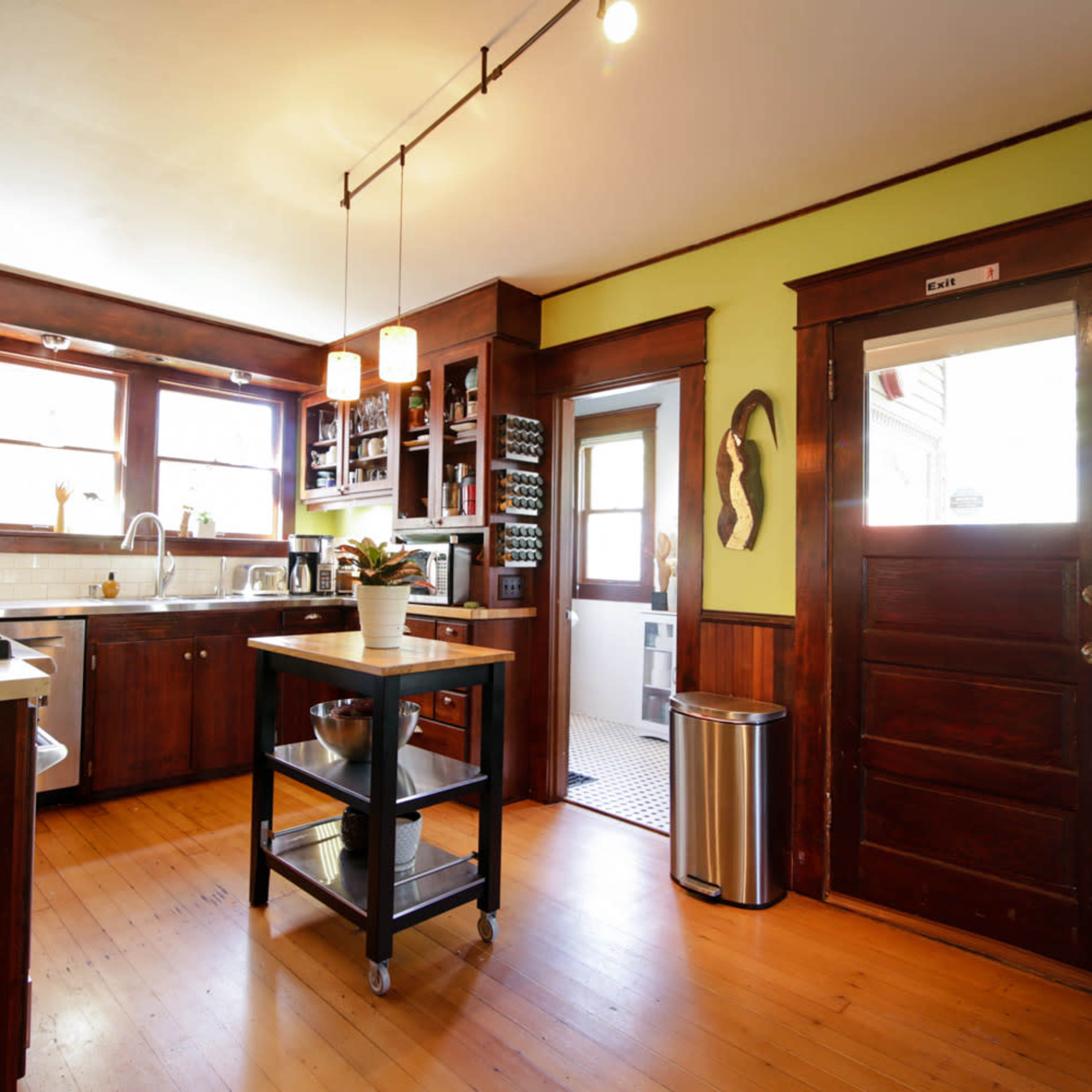 A kitchen features wooden cabinets, a cooking range, a central island on wheels, and a door leading to an adjacent room.