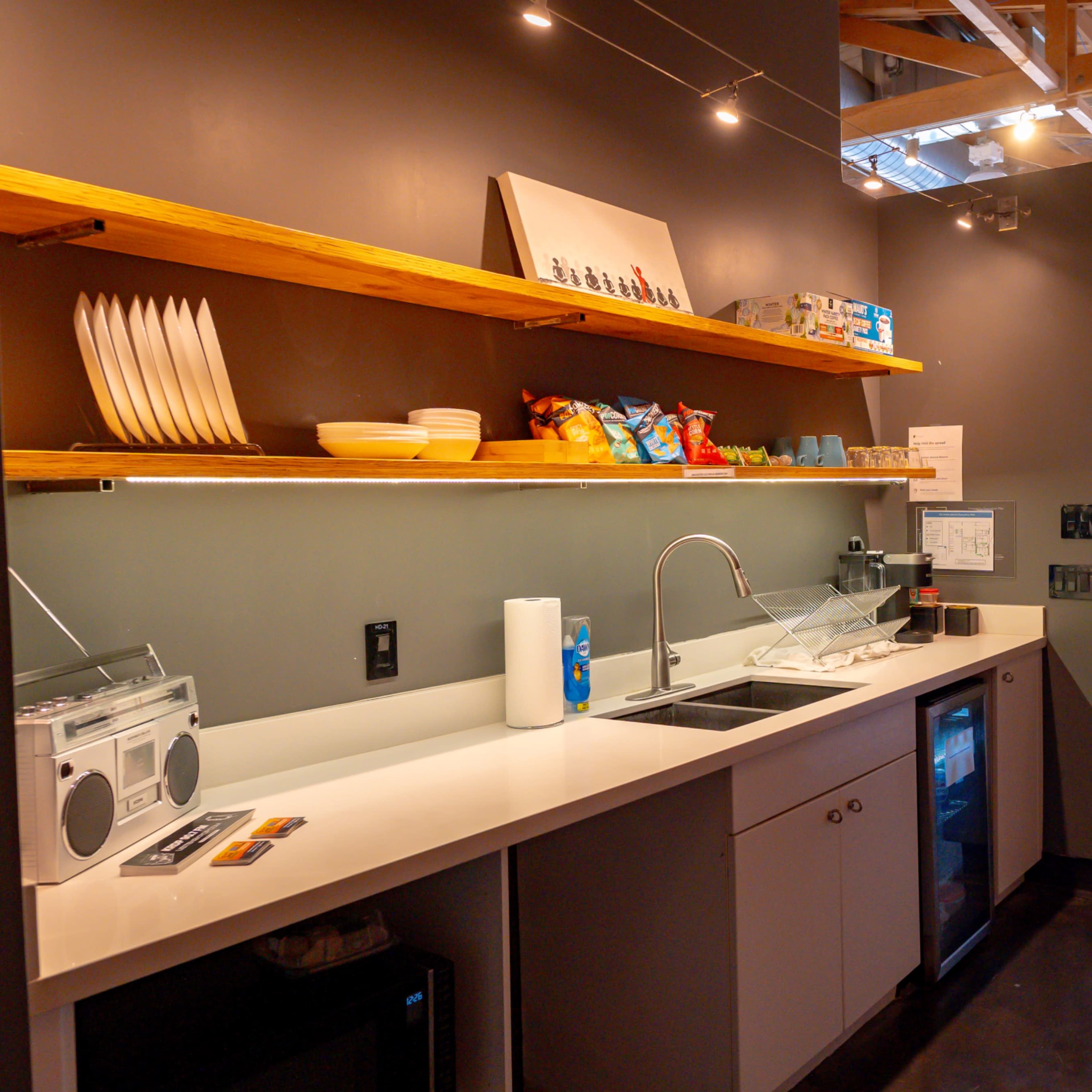 A small kitchen area features a counter with a sink, shelves holding dishes and snacks, and a mini fridge, all set against a sleek gray wall.