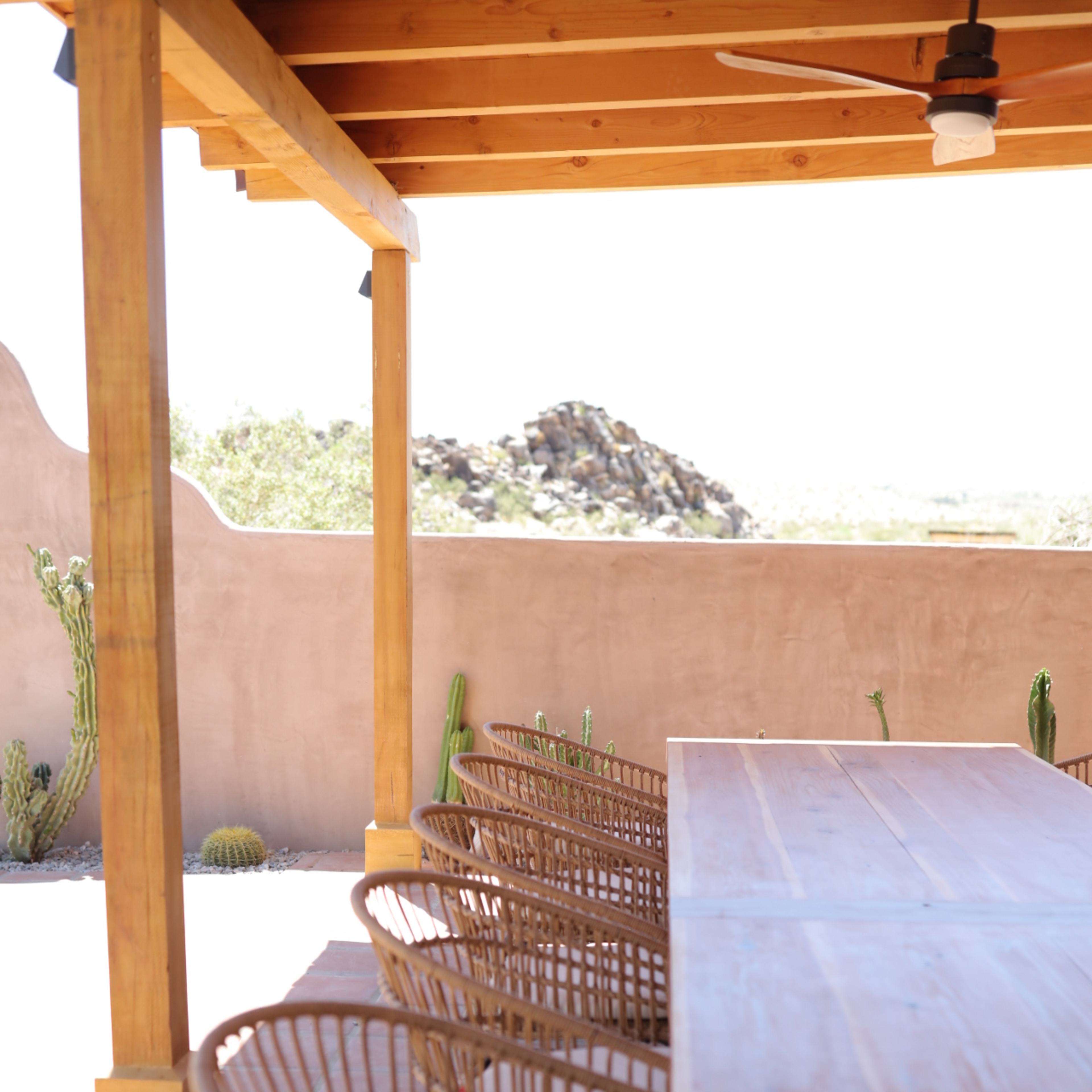 A wooden table with several wicker chairs is positioned under a wooden pergola, overlooking a desert landscape with cacti and rocky hills in the background.