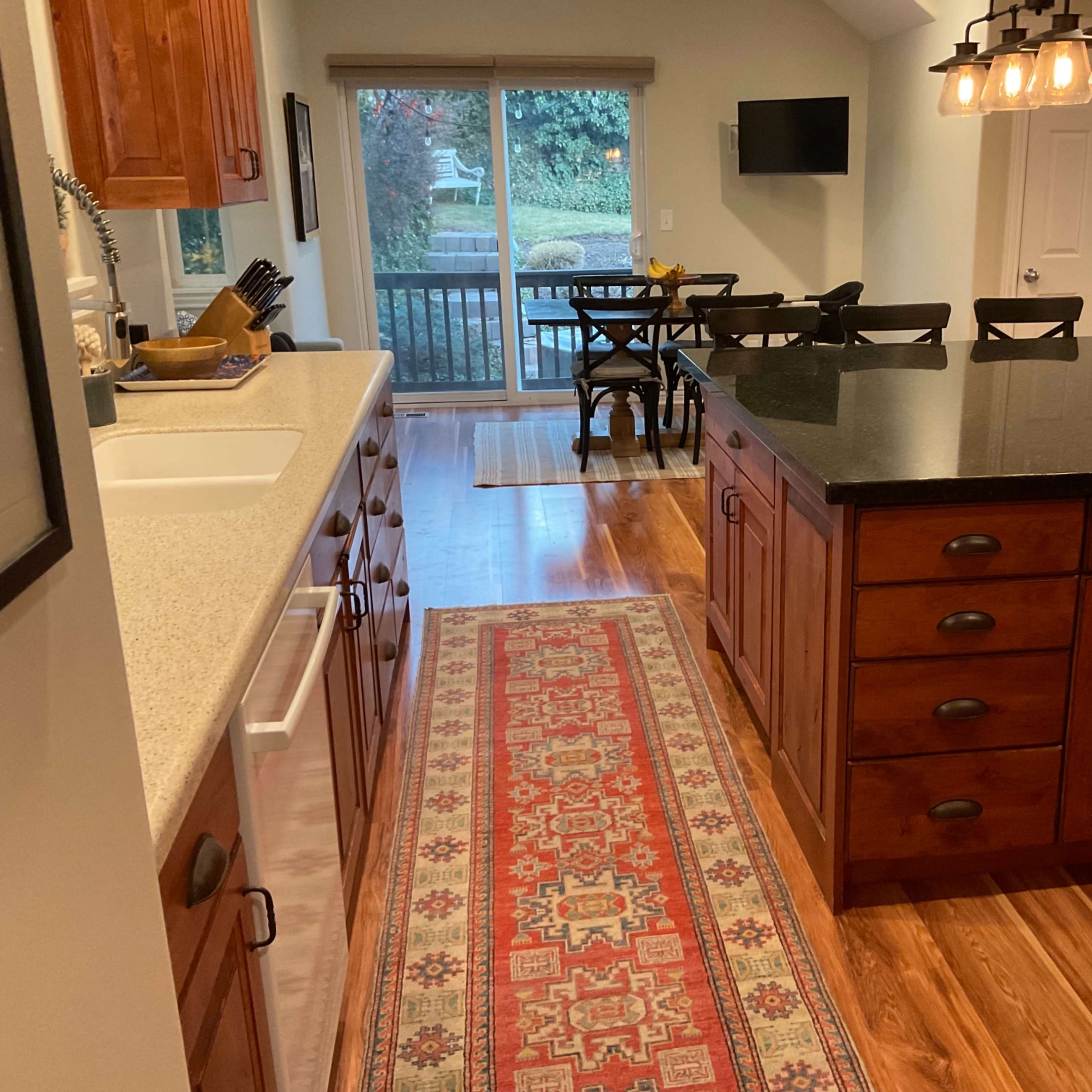 A modern kitchen features wooden cabinets, a large island with a black countertop, and a dining area visible through the sliding glass door.