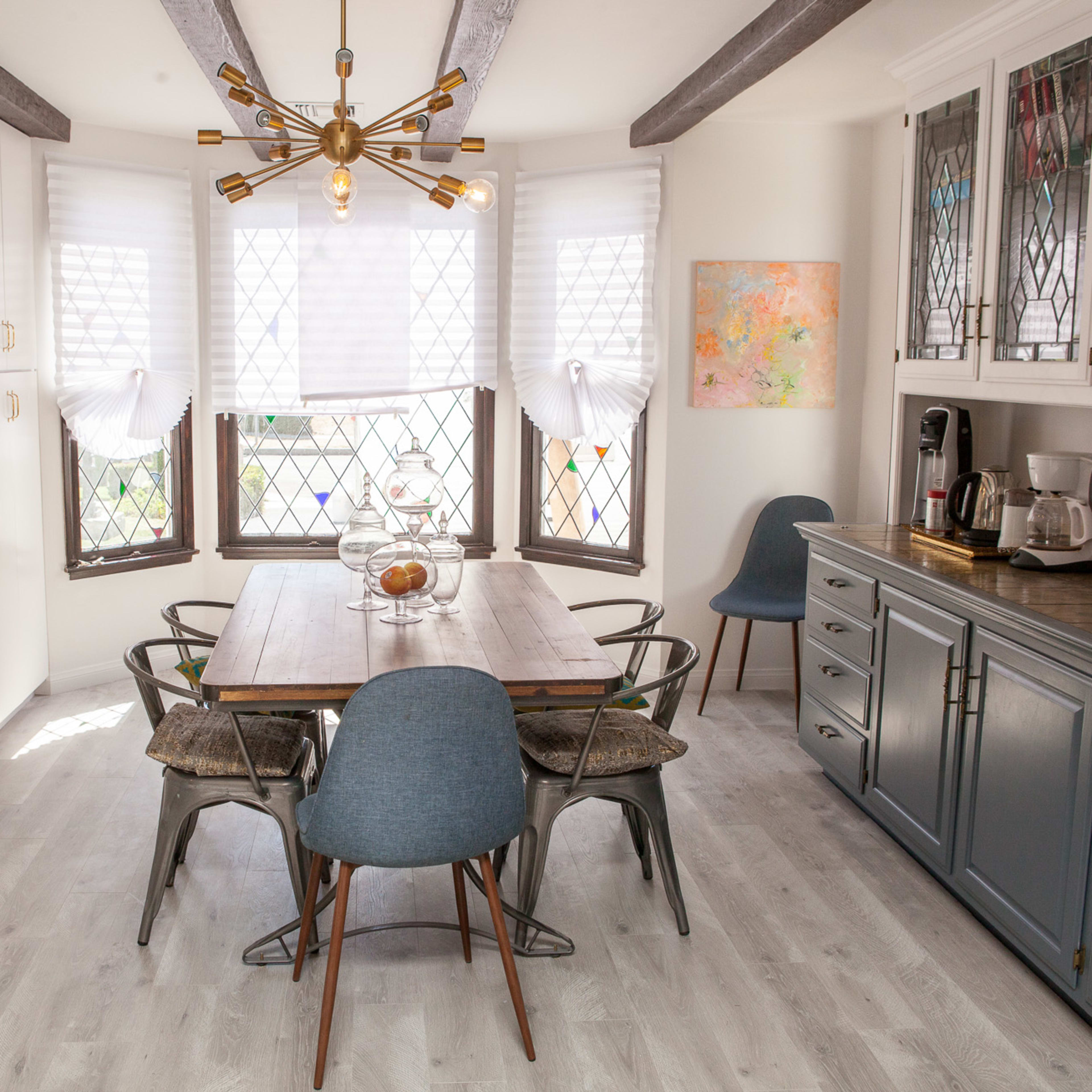 A bright kitchen features a wooden table surrounded by chairs, with large windows adorned with shades and a modern light fixture above.