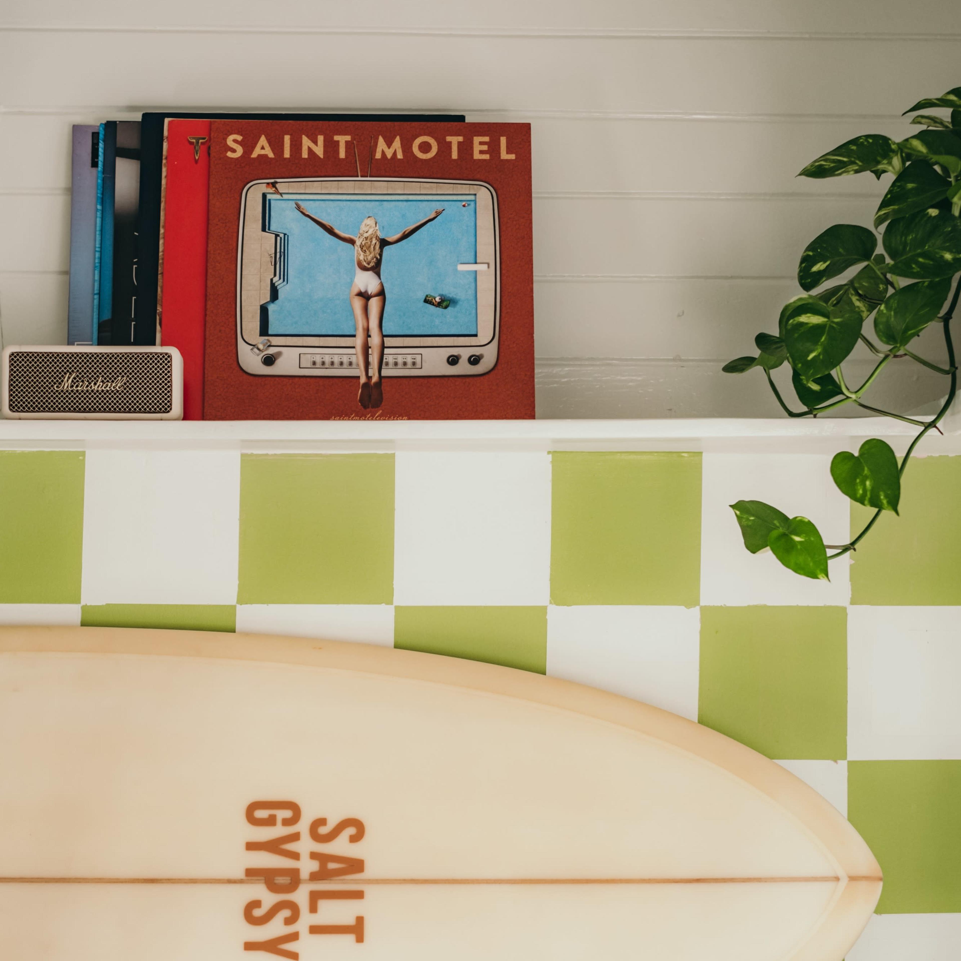 A surfboard labeled "SALT GYPSY" rests on a green and white checkered surface beneath a stack of vinyl records and a potted plant.