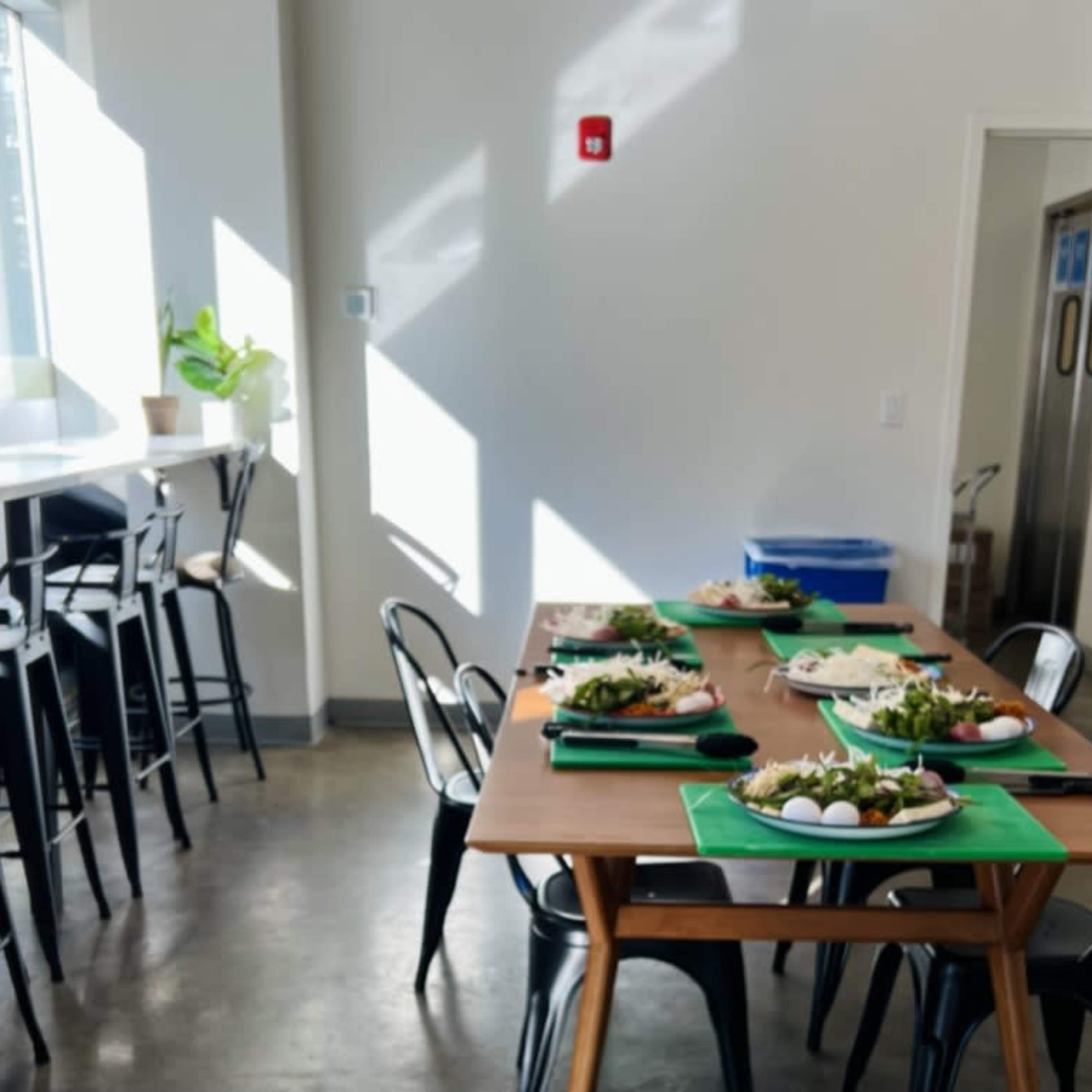A dining table with multiple plates of food is set up in a brightly lit room featuring high chairs and a plant.