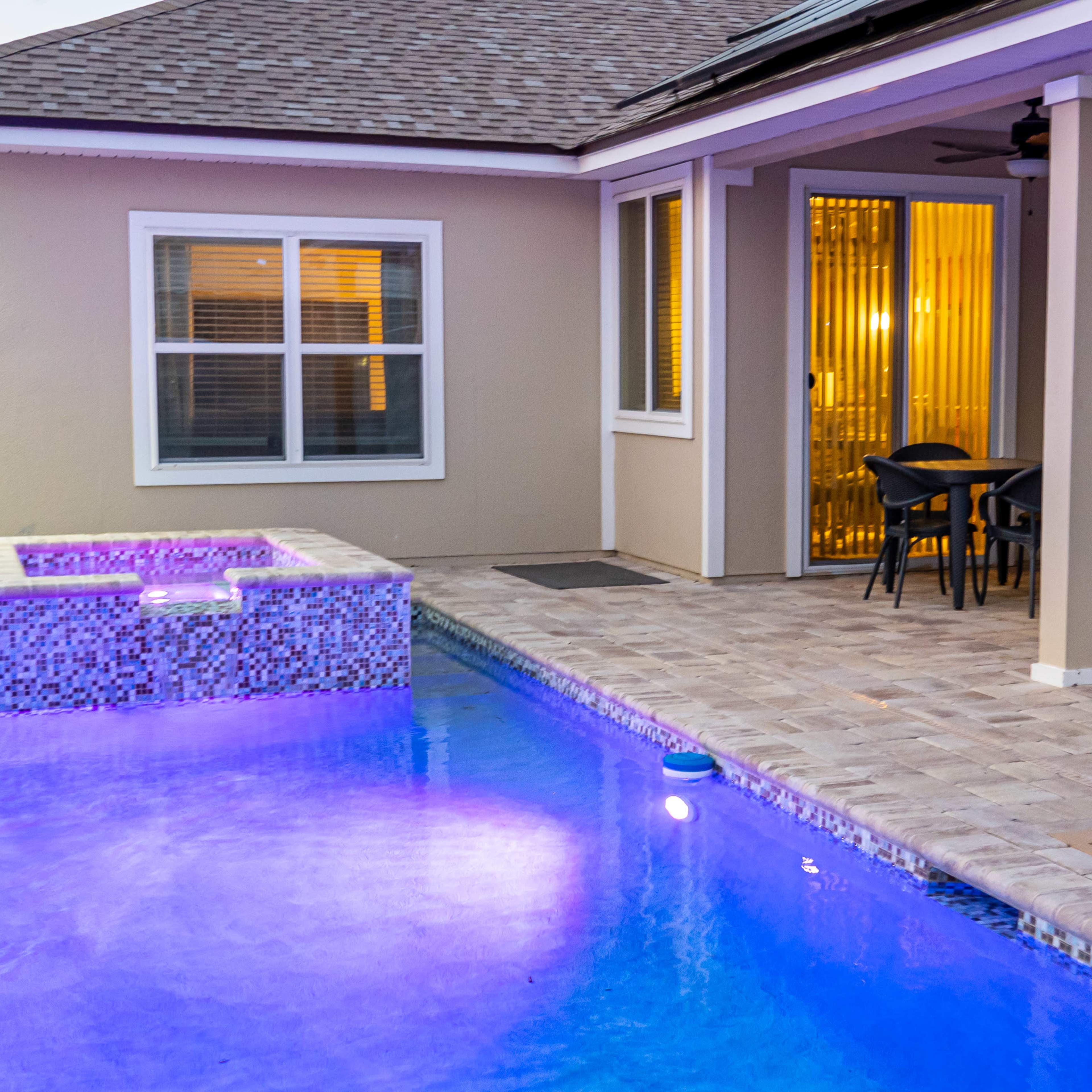 A backyard area with a swimming pool featuring a spa, surrounded by tiled flooring and a set of outdoor furniture under a covered patio.
