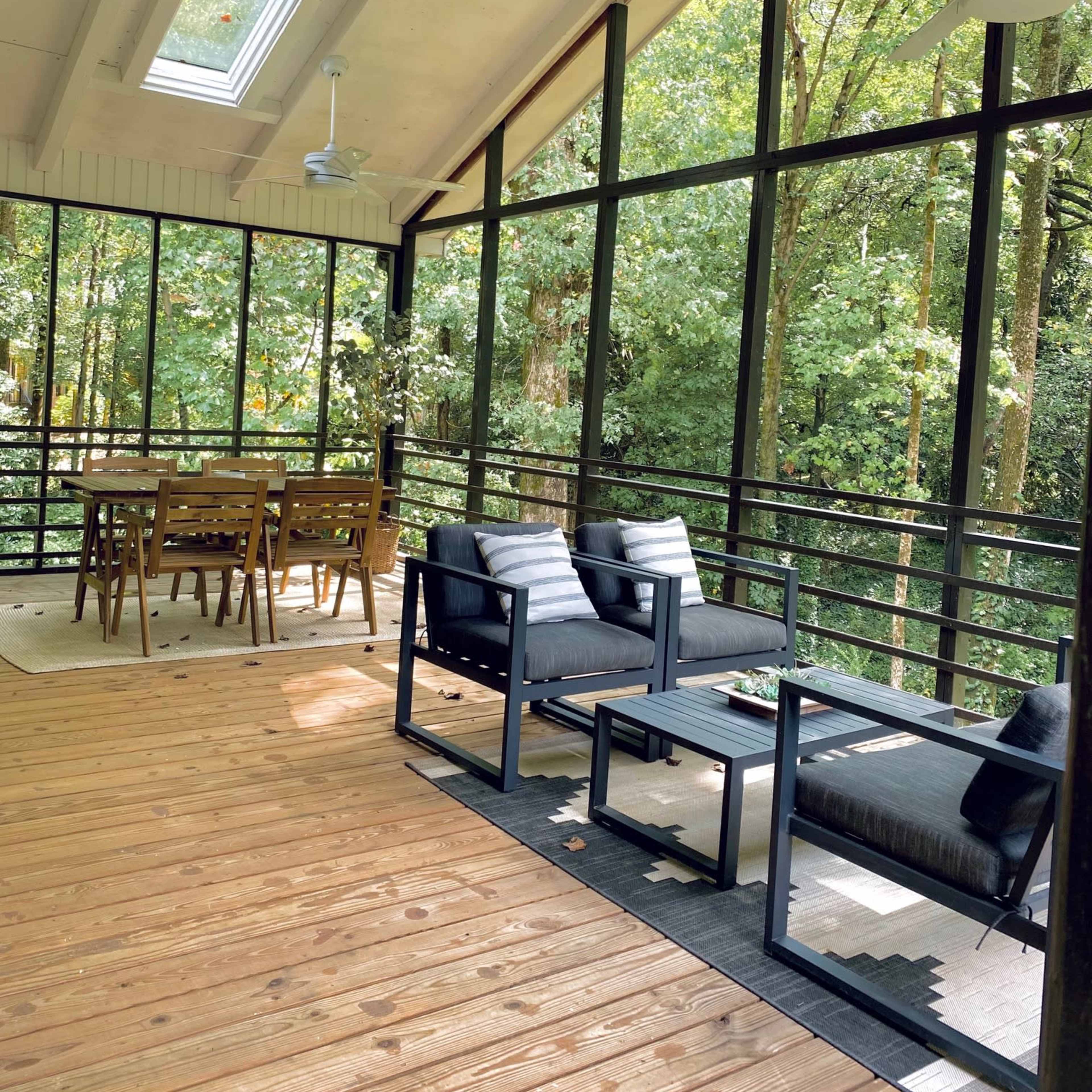 A screened-in porch with wooden flooring, two modern seating areas, a dining table, and large windows overlooking a wooded area.