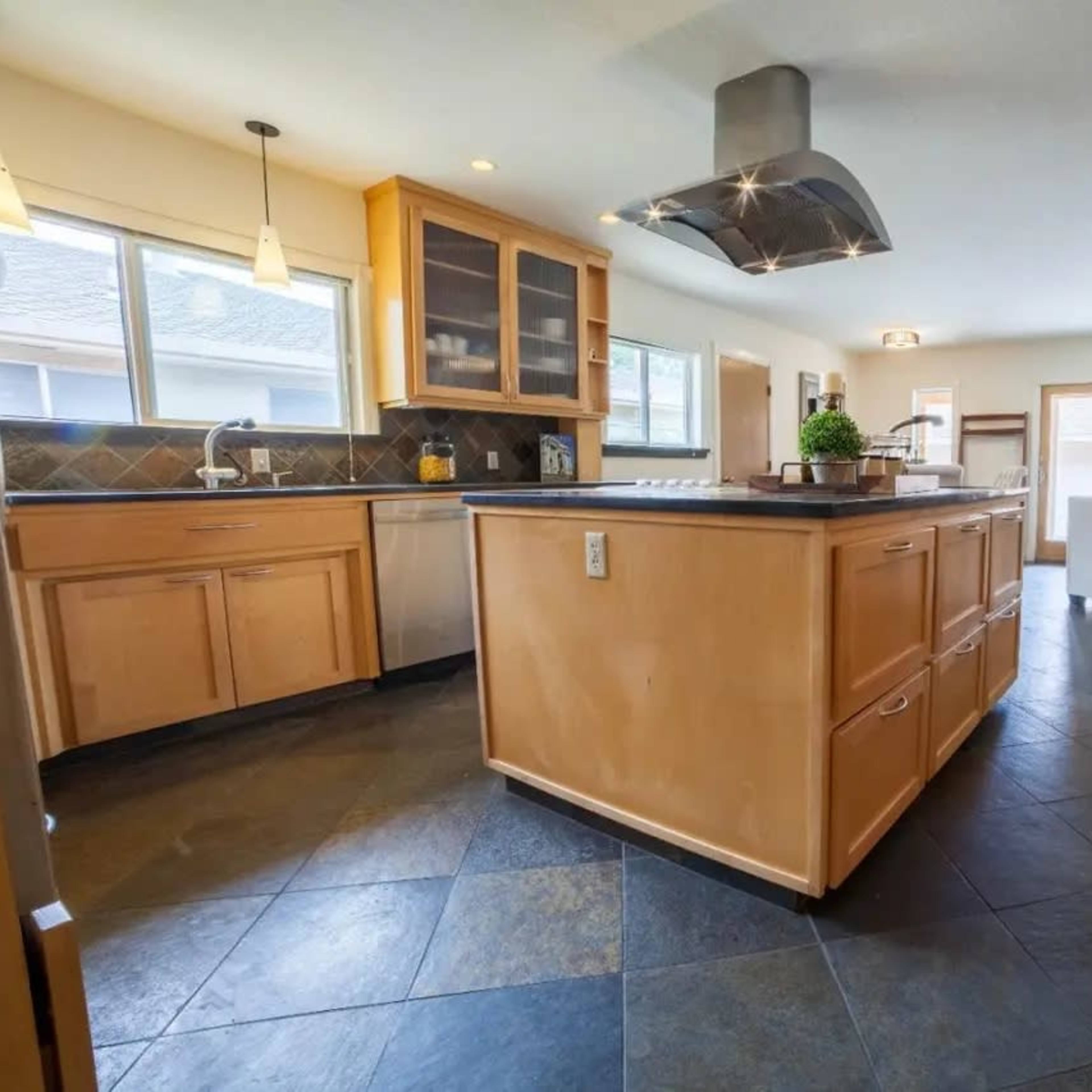 The kitchen features wooden cabinetry, a large central island, and a slate tile floor, with windows providing natural light.