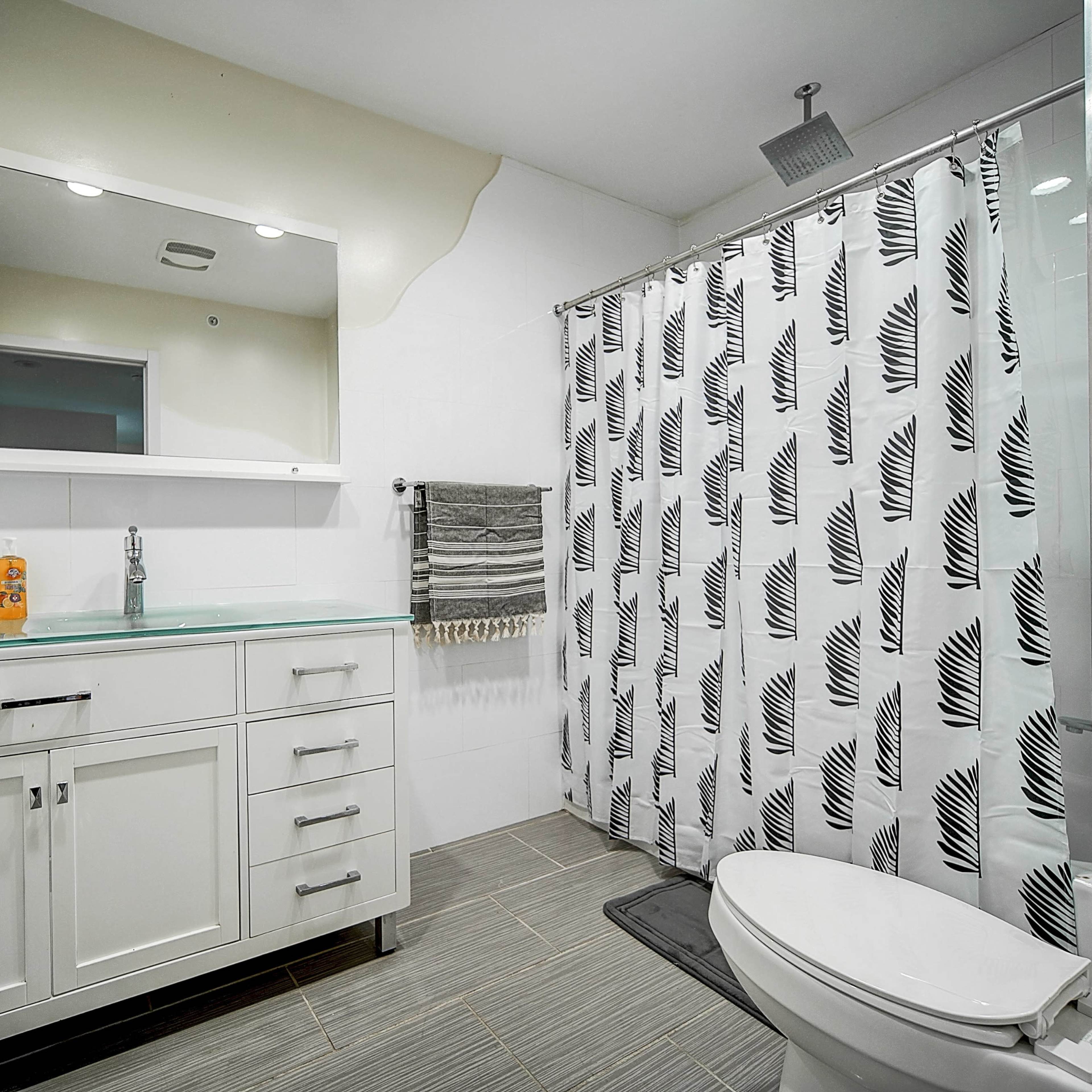 A modern bathroom with a white vanity, a mirror, a shower curtain featuring a leaf pattern, and a toilet.