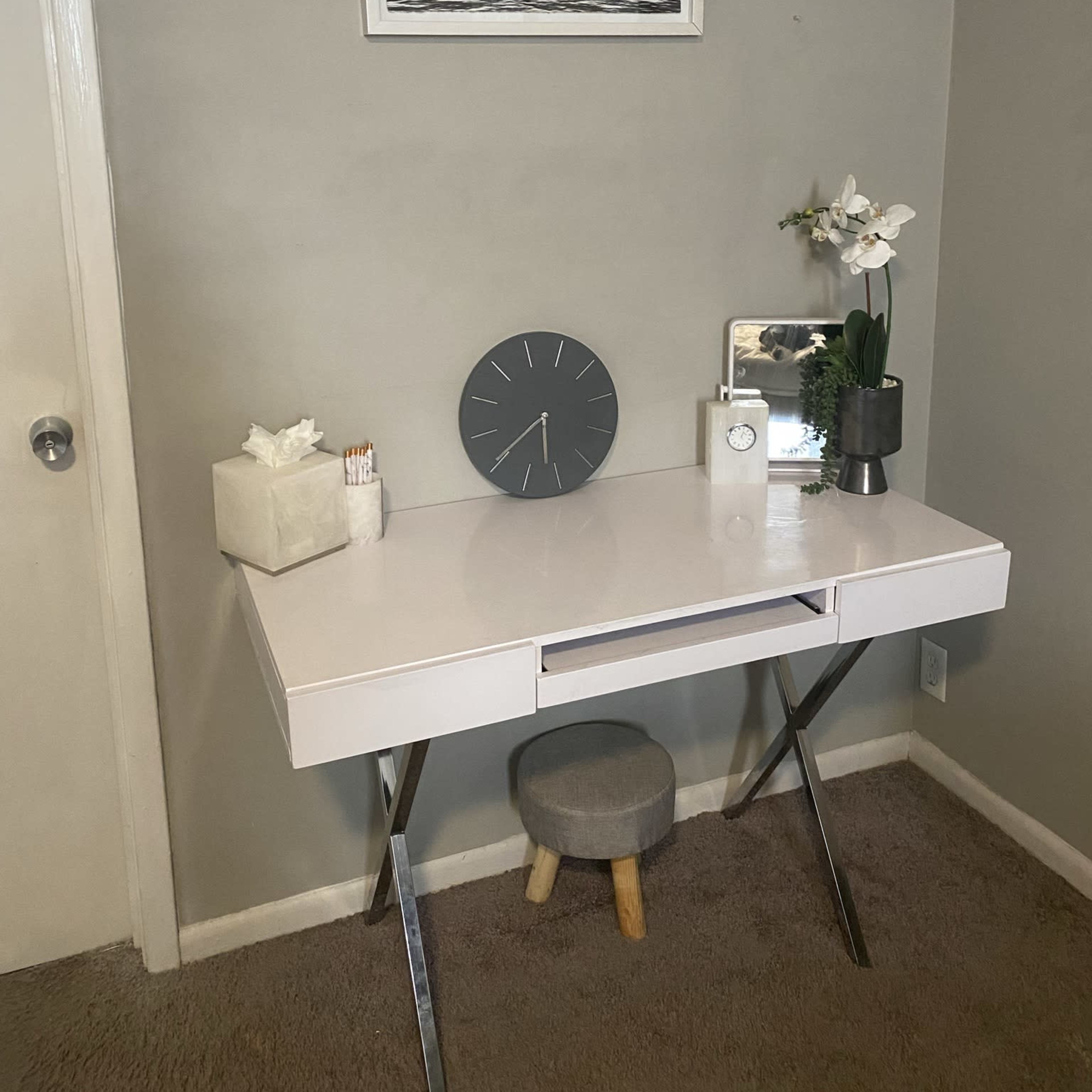 A minimalistic white desk with a drawer, a round clock, a mirror, a vase with flowers, and a small stool against a gray wall.