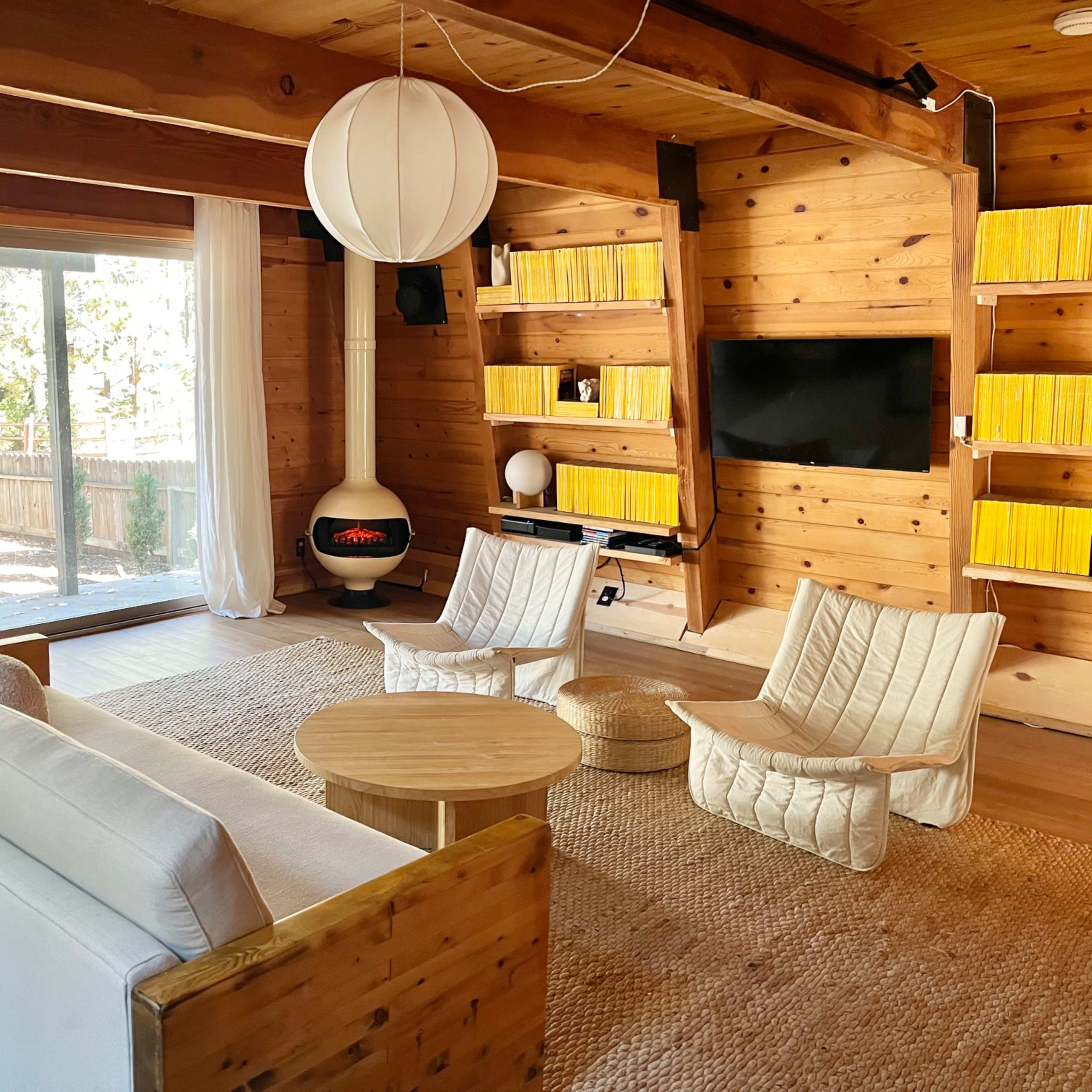 A modern wooden living room with a round coffee table, two armchairs, a sofa, and a fireplace, all arranged near large windows that let in natural light.