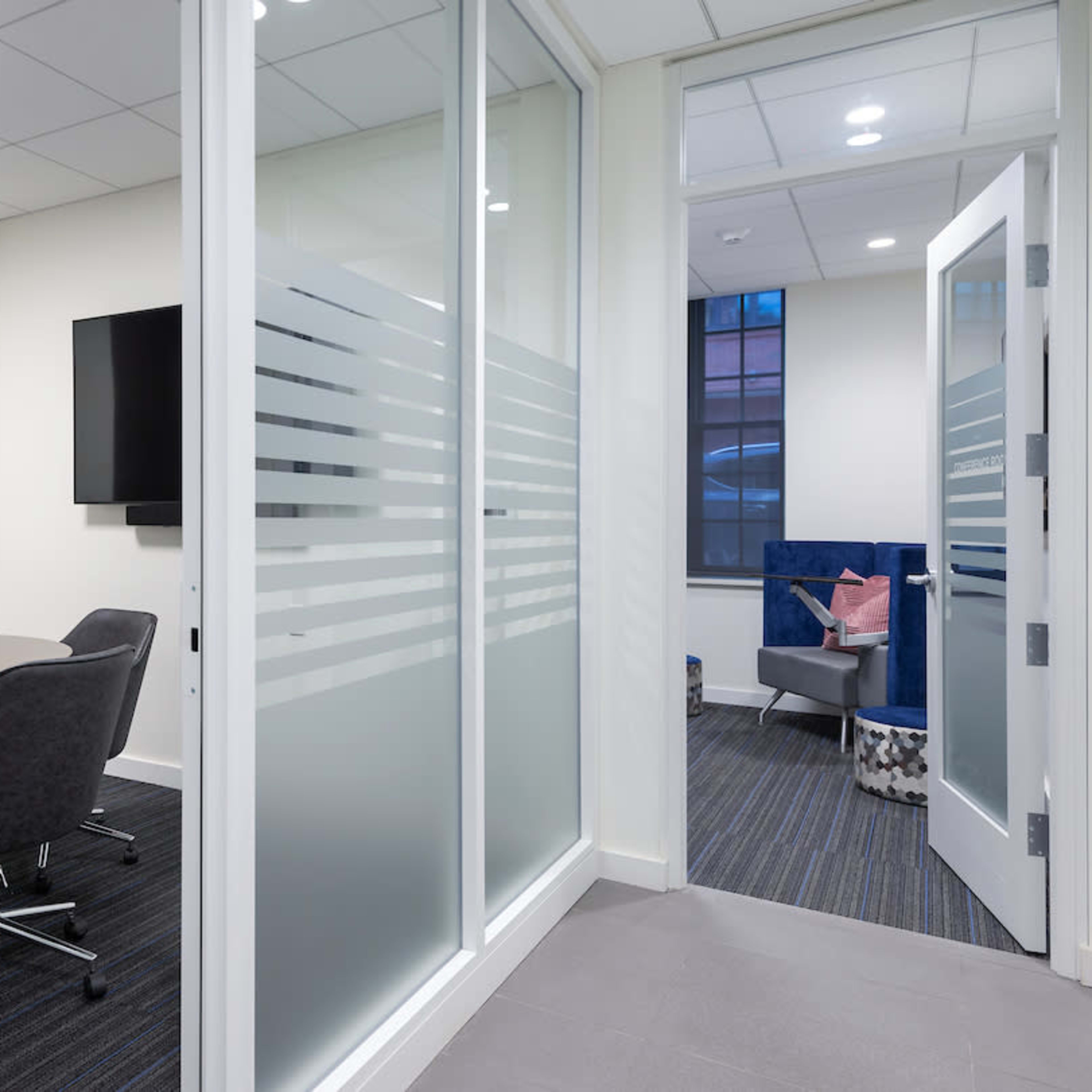 A modern office space with a conference room featuring a round table and chairs, adjacent to a lounge area visible through an open door.
