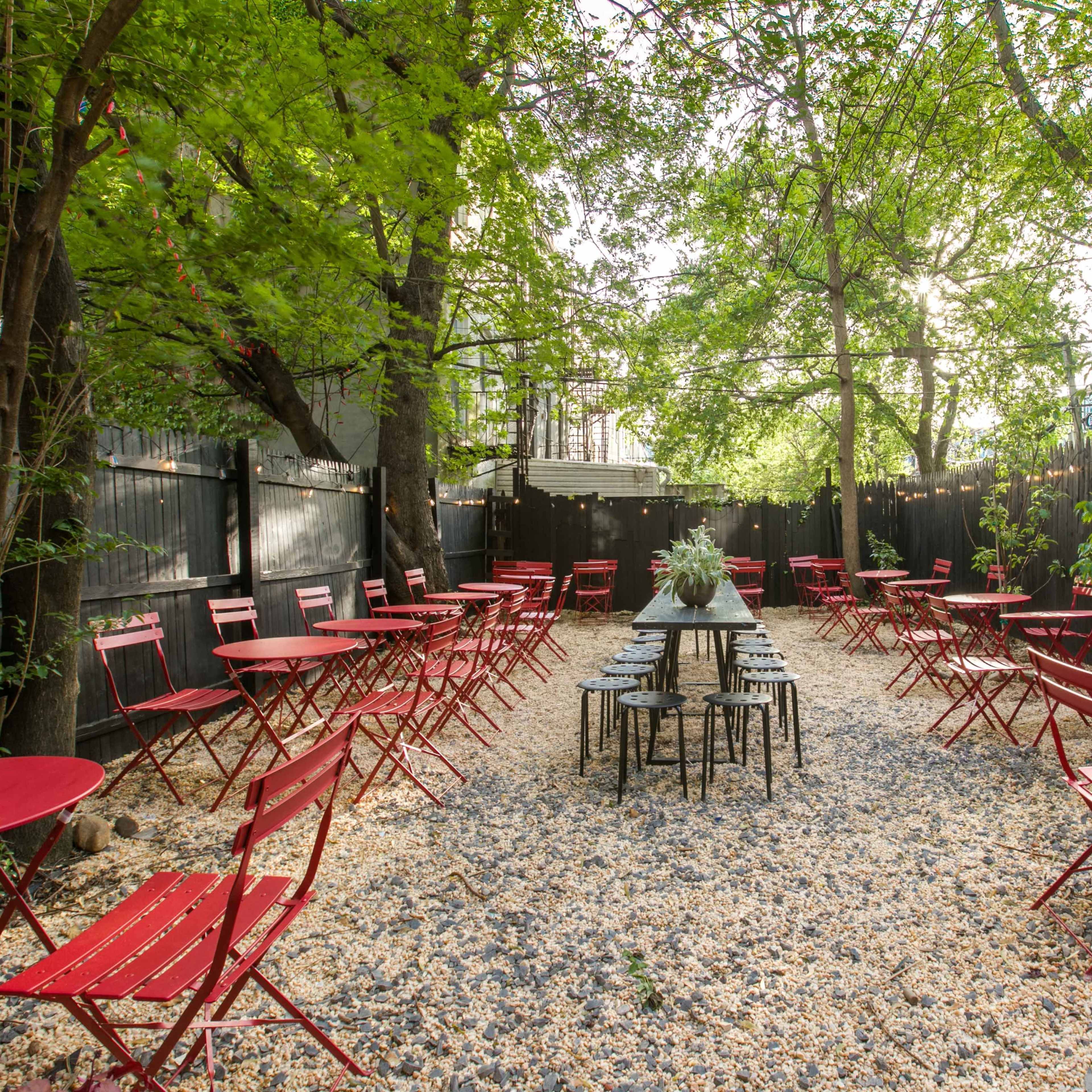 A garden café features red metal tables and chairs arranged on a gravel path surrounded by trees.