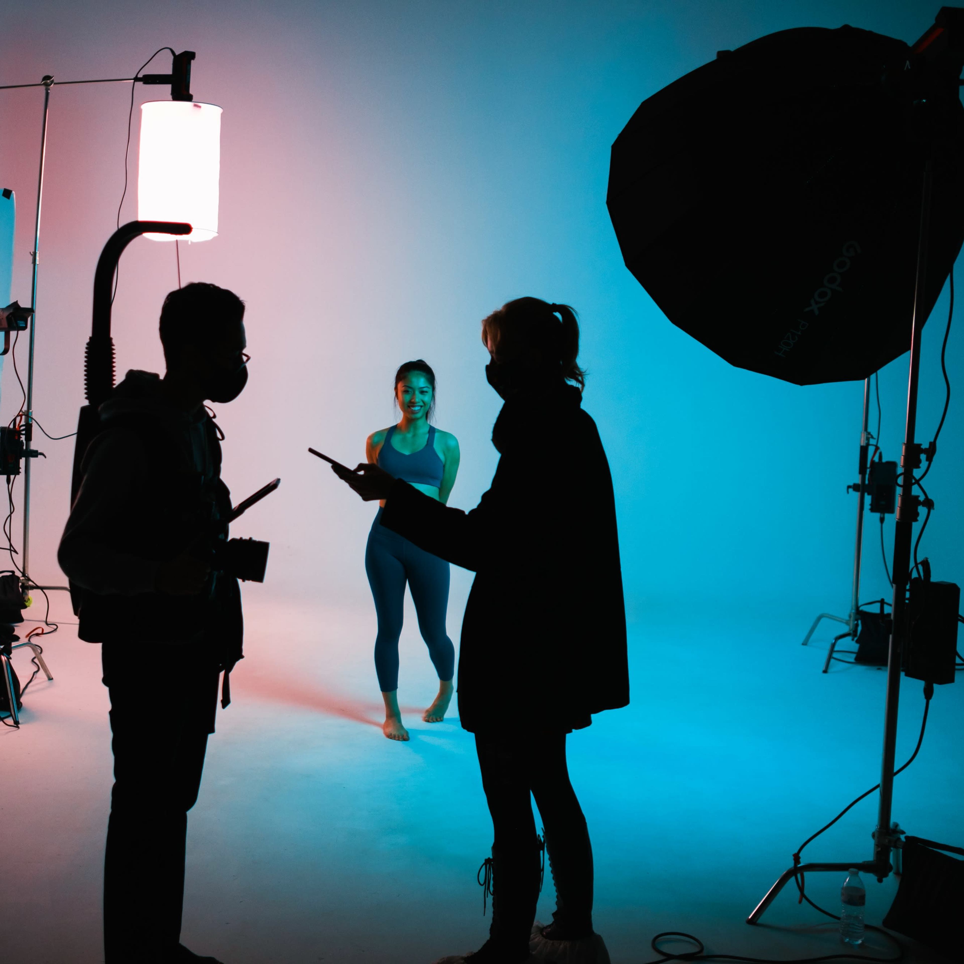 A dancer poses under studio lights while two crew members discuss setup in the foreground.