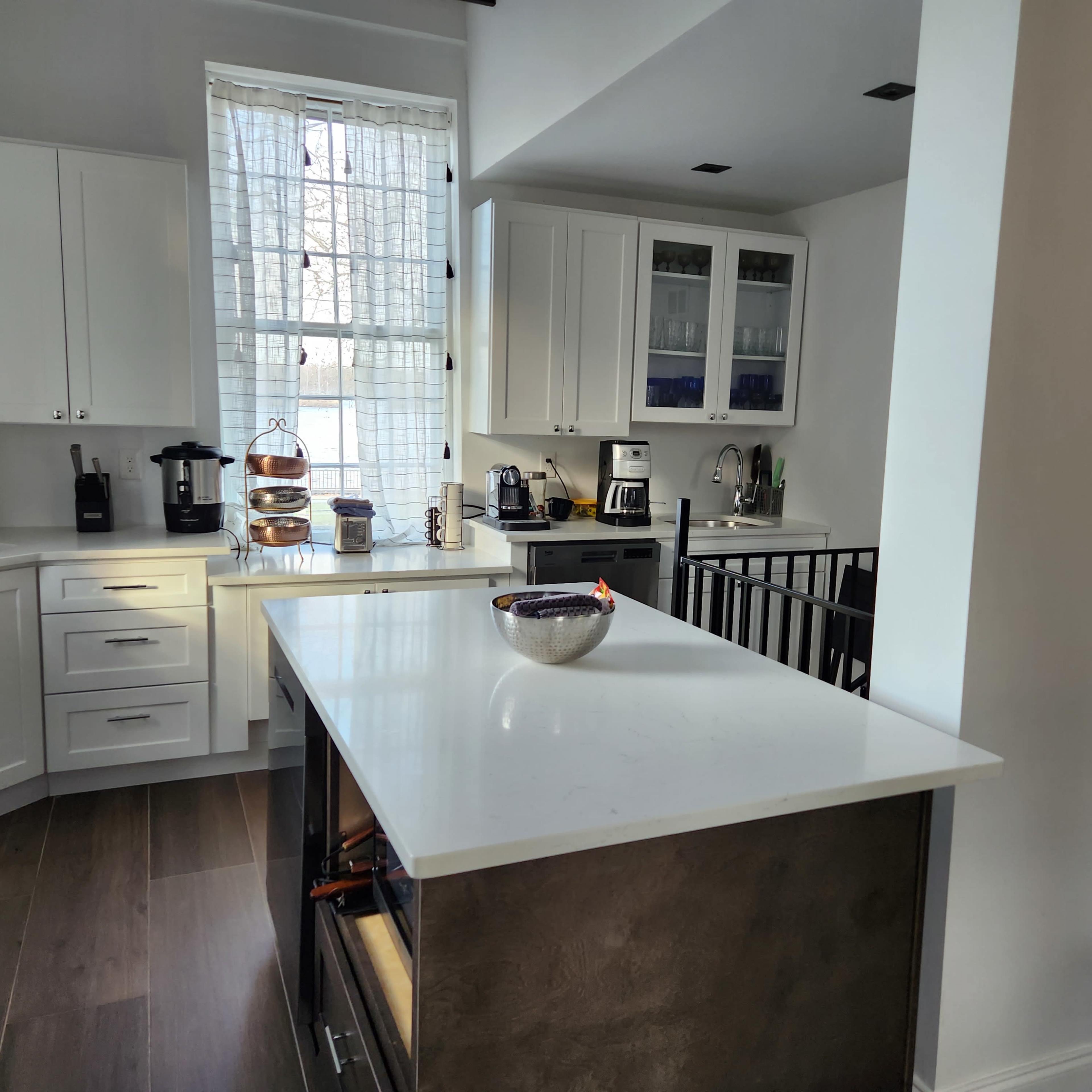 A modern kitchen features white cabinets, a central island, and various appliances, with a window allowing natural light to fill the space.