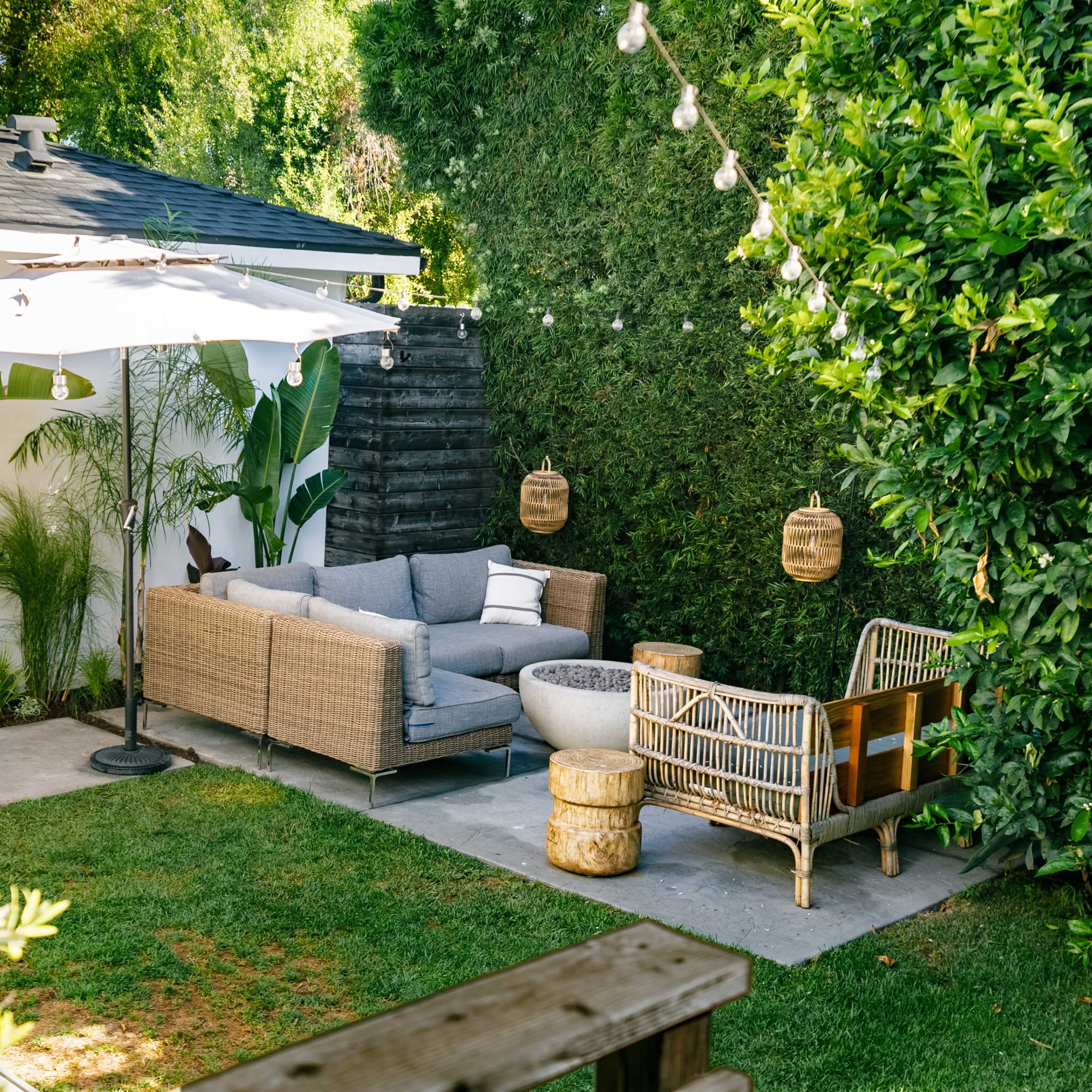 A patio area with a wicker sofa, a sun umbrella, a small table, and two decorative chairs surrounded by greenery.