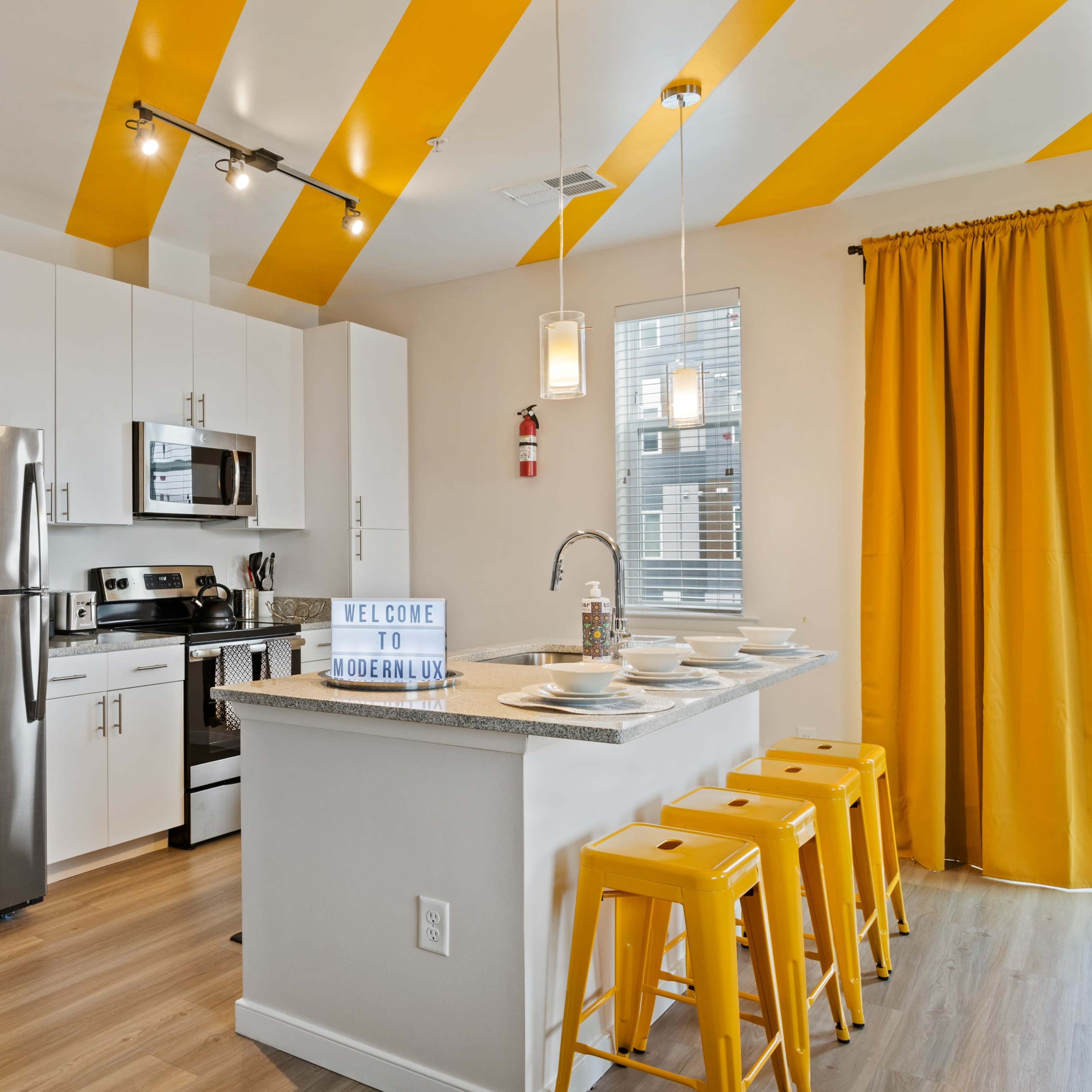 A modern kitchen features stainless steel appliances, yellow accents, and a breakfast bar with yellow stools.