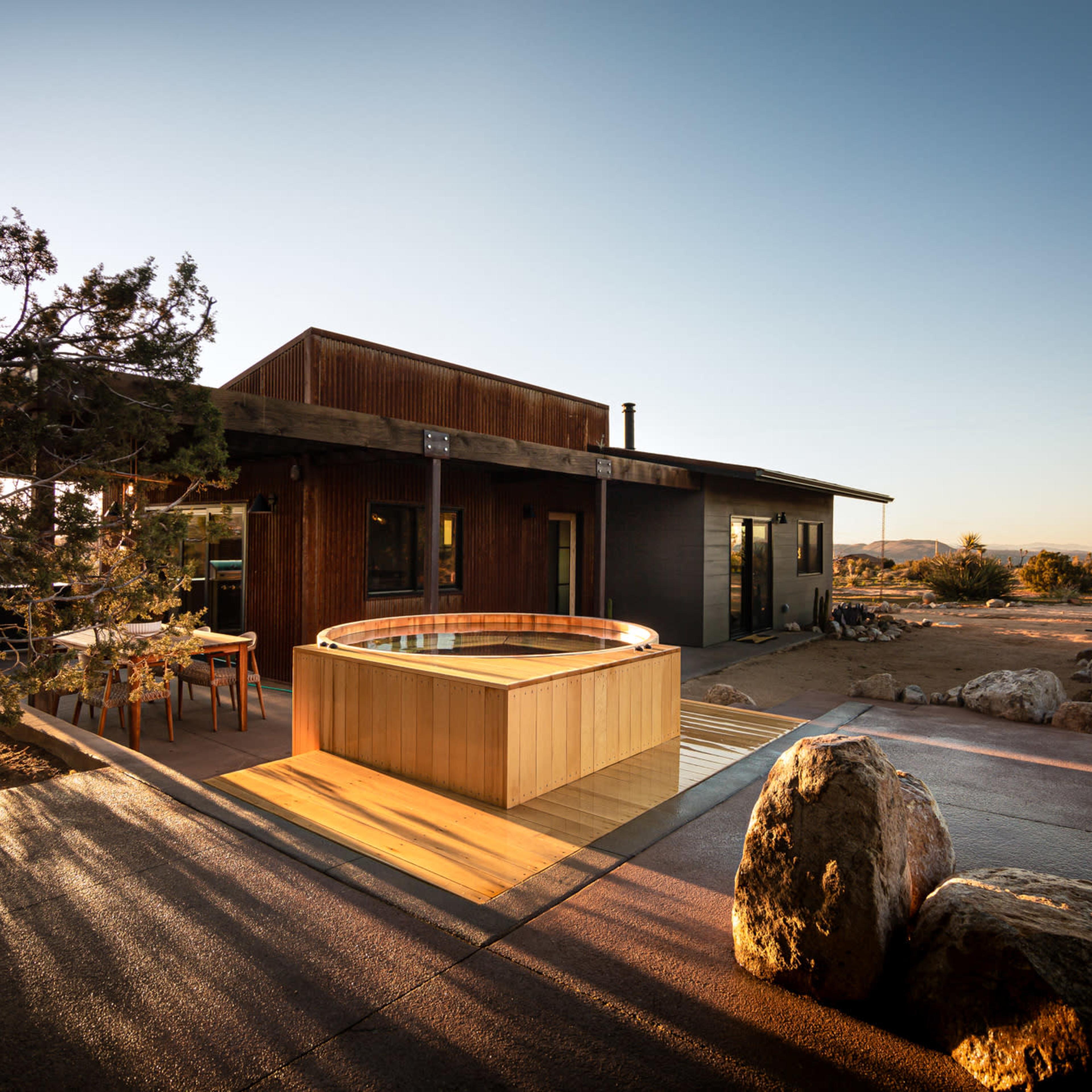 A modern cabin features a wooden hot tub on a patio surrounded by rocks and desert vegetation.