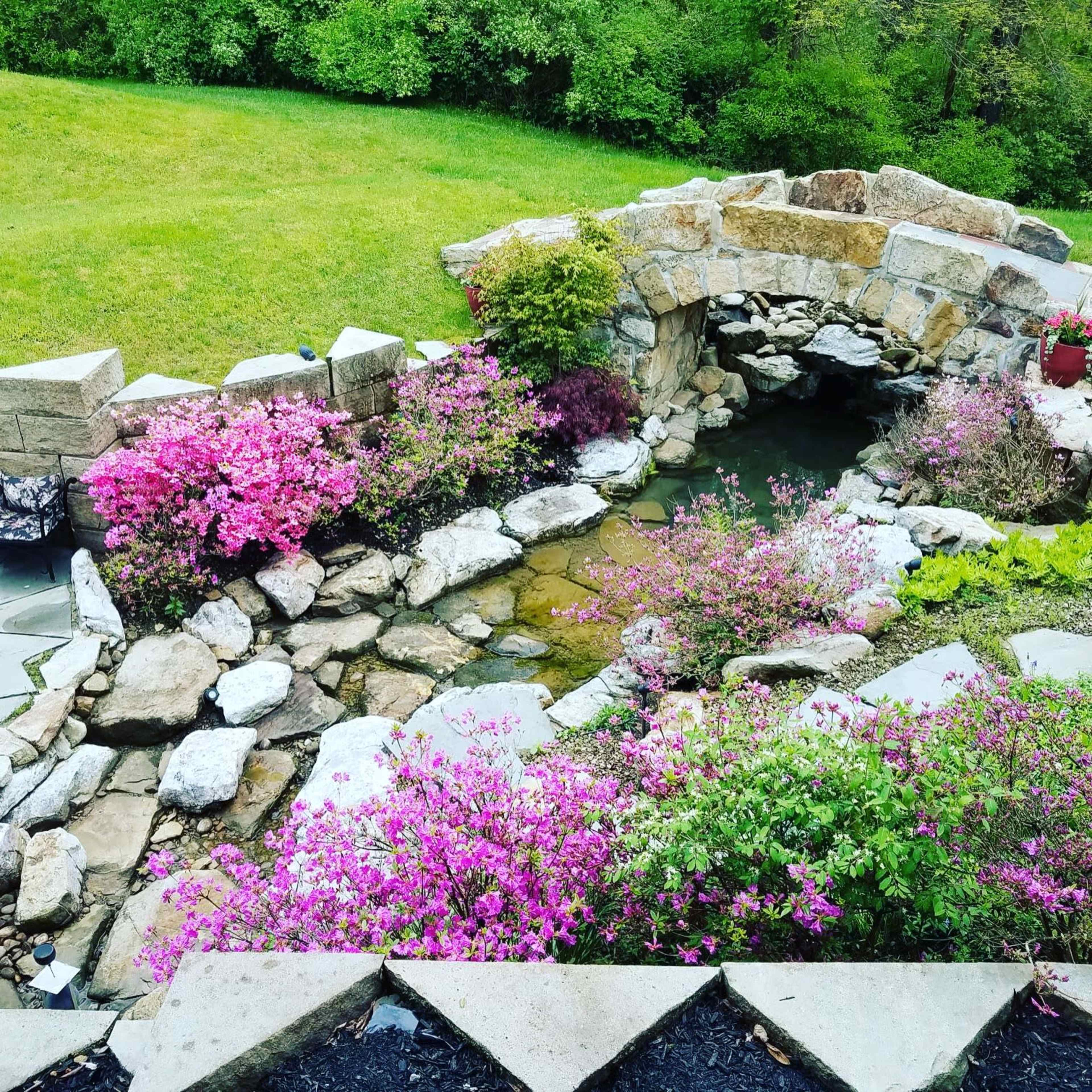 A stone bridge arching over a pond surrounded by flowering plants and greenery.