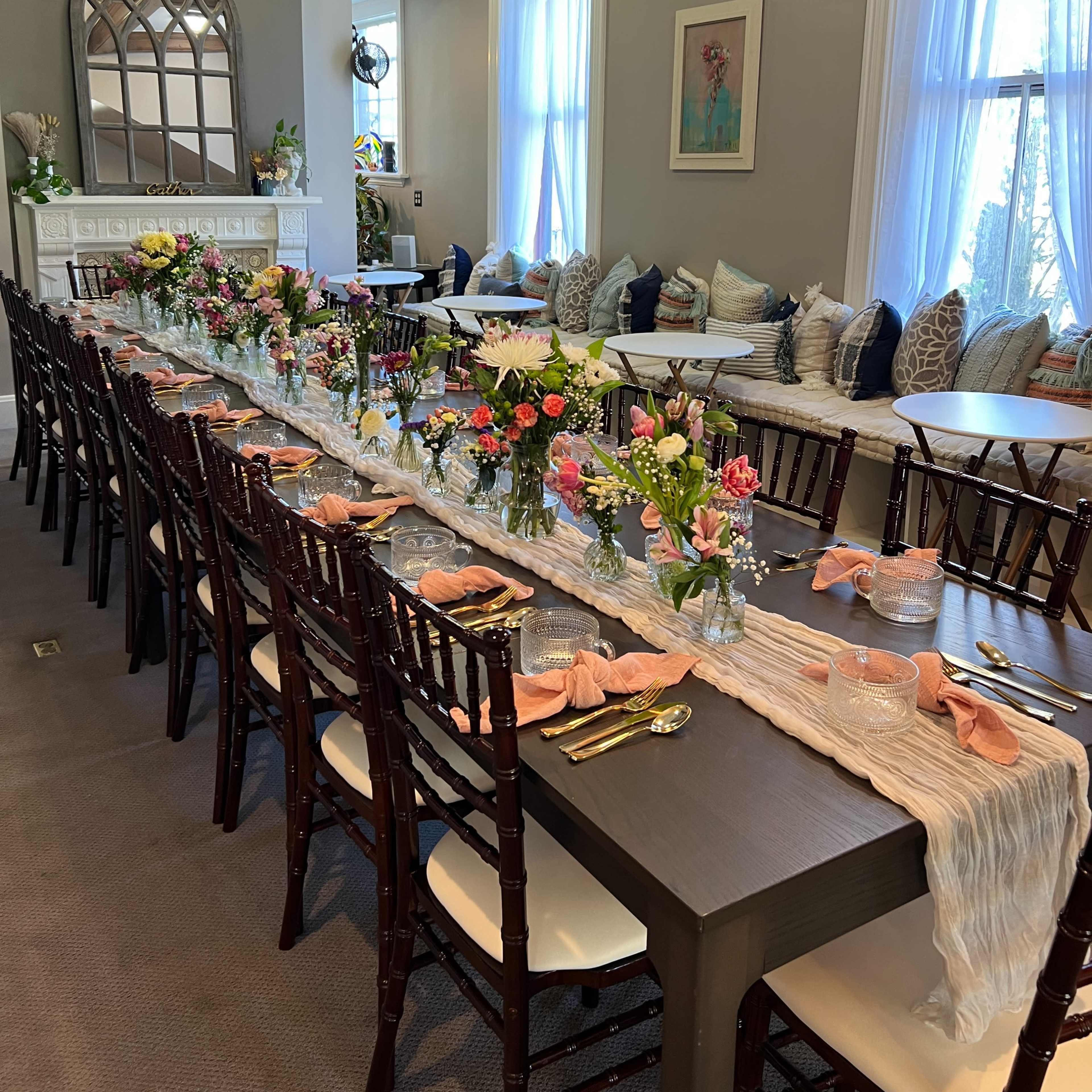 A long dining table is set with floral arrangements and decorative tableware in a brightly lit room featuring a fireplace and cushioned seating.