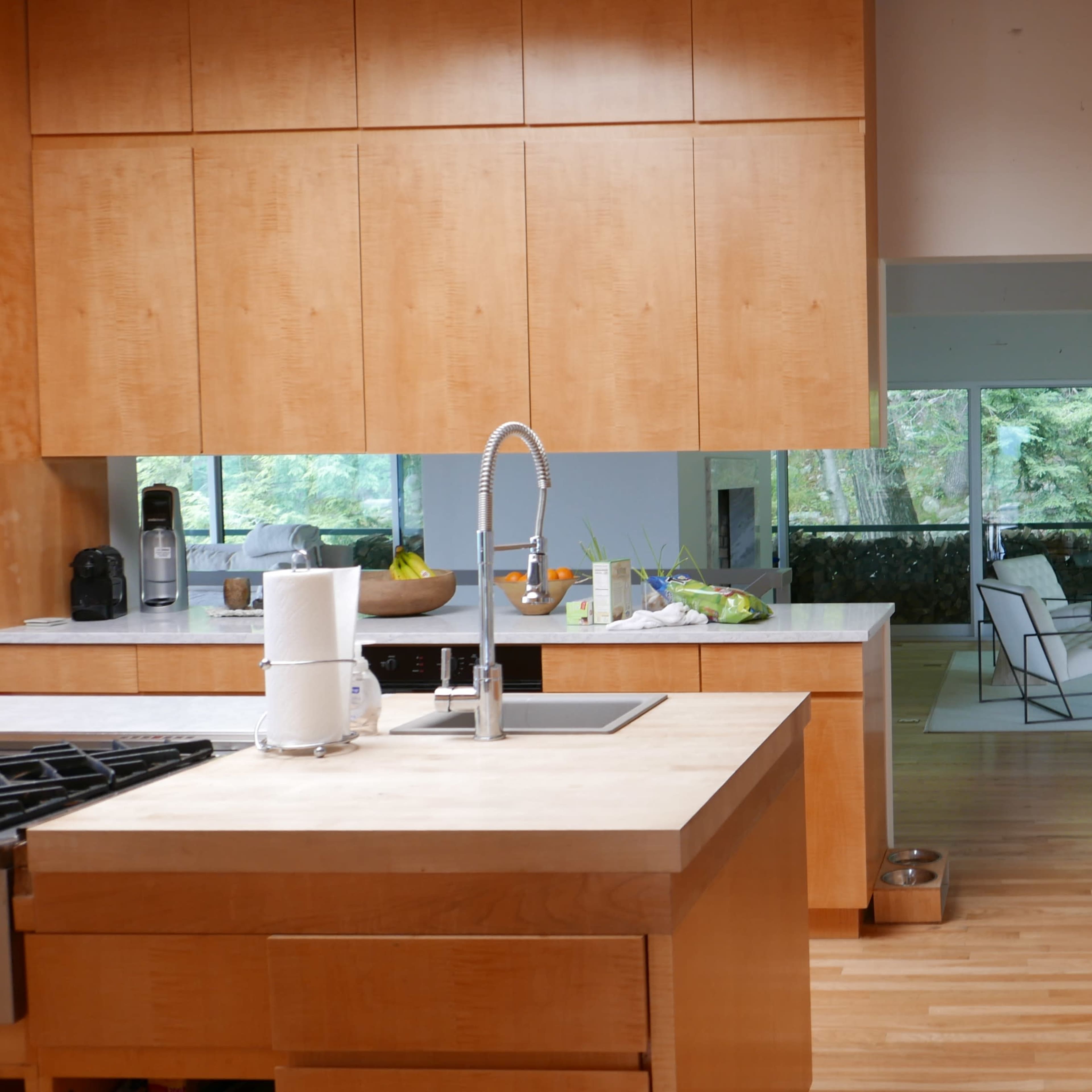 A modern kitchen with wooden cabinetry, a central island with a sink, and a view into a living area with large windows.