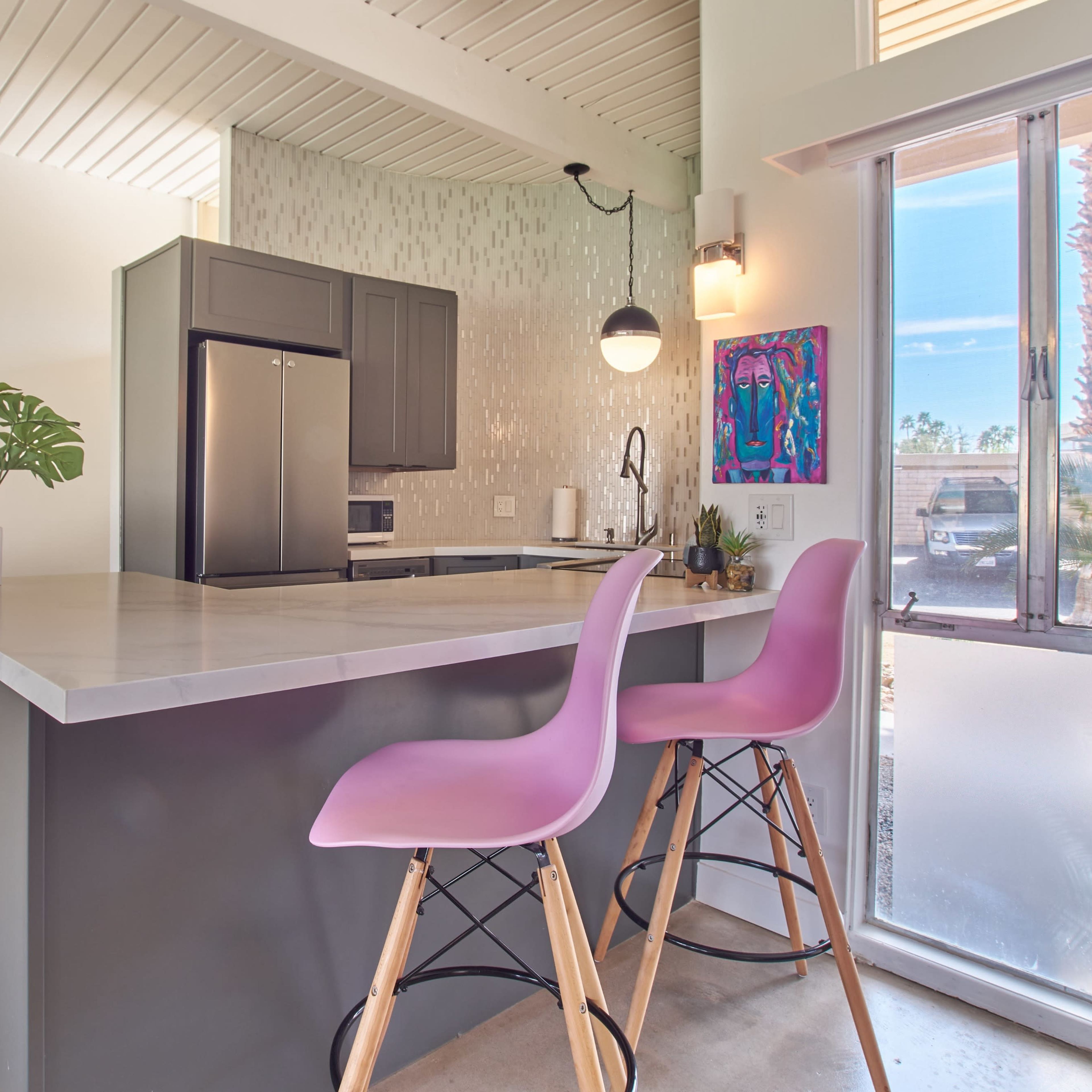 A modern kitchen features a countertop with two pink stools, stainless steel appliances, and a large window providing natural light.