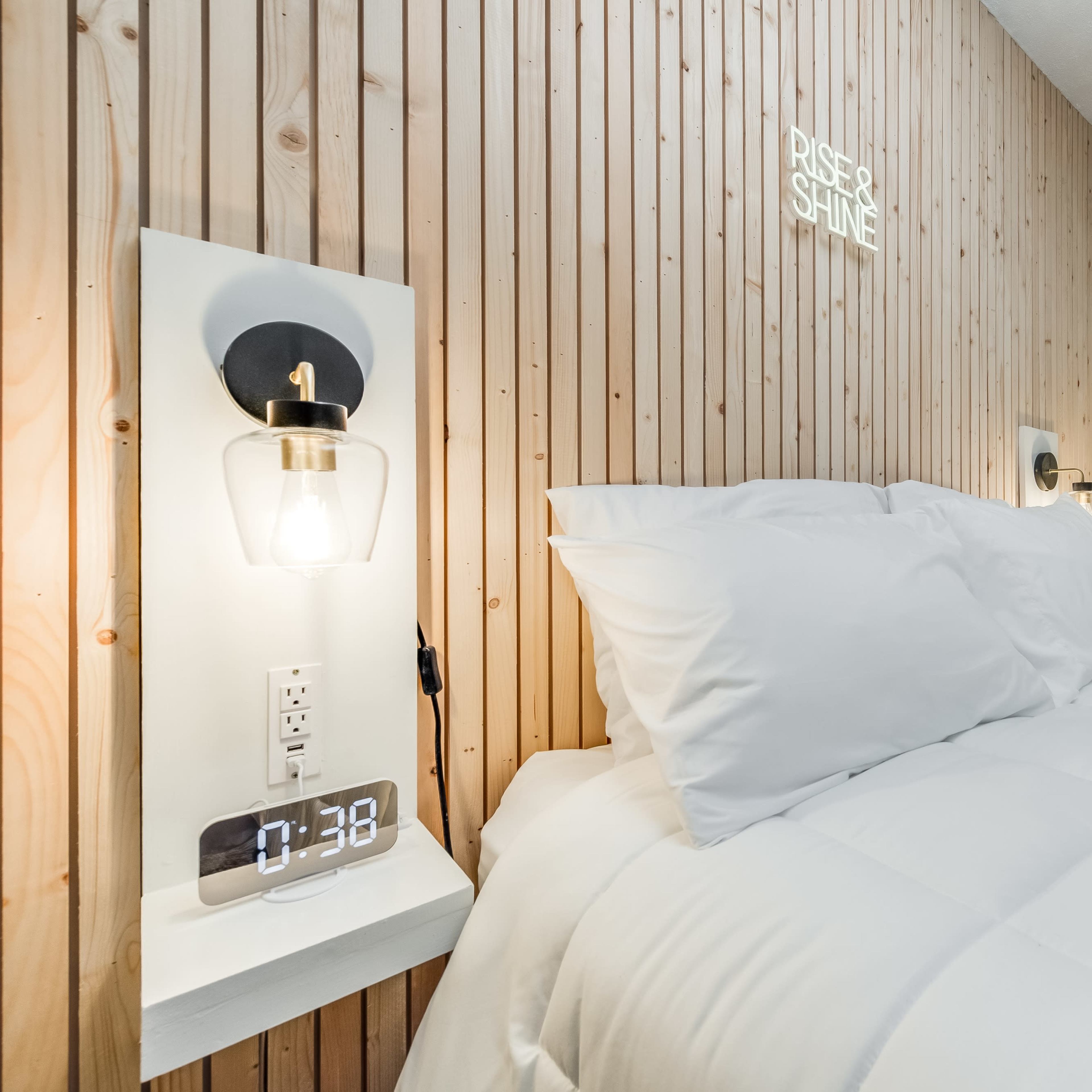 A neatly made bed with white bedding is positioned against a wooden-paneled wall, featuring minimalist bedside lamps and a clock.
