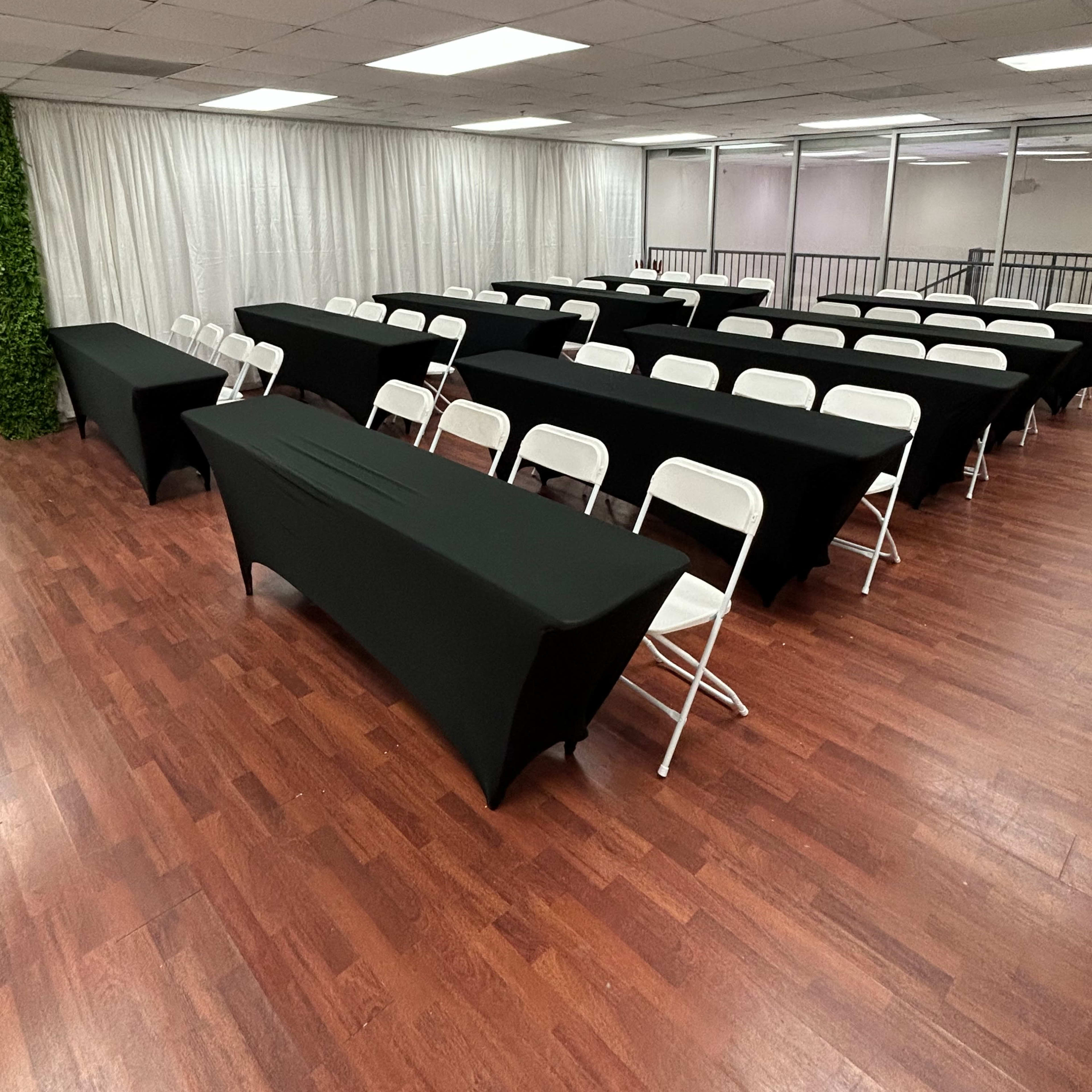 A spacious indoor venue features several rows of black-draped tables and white folding chairs arranged neatly on a wooden floor.