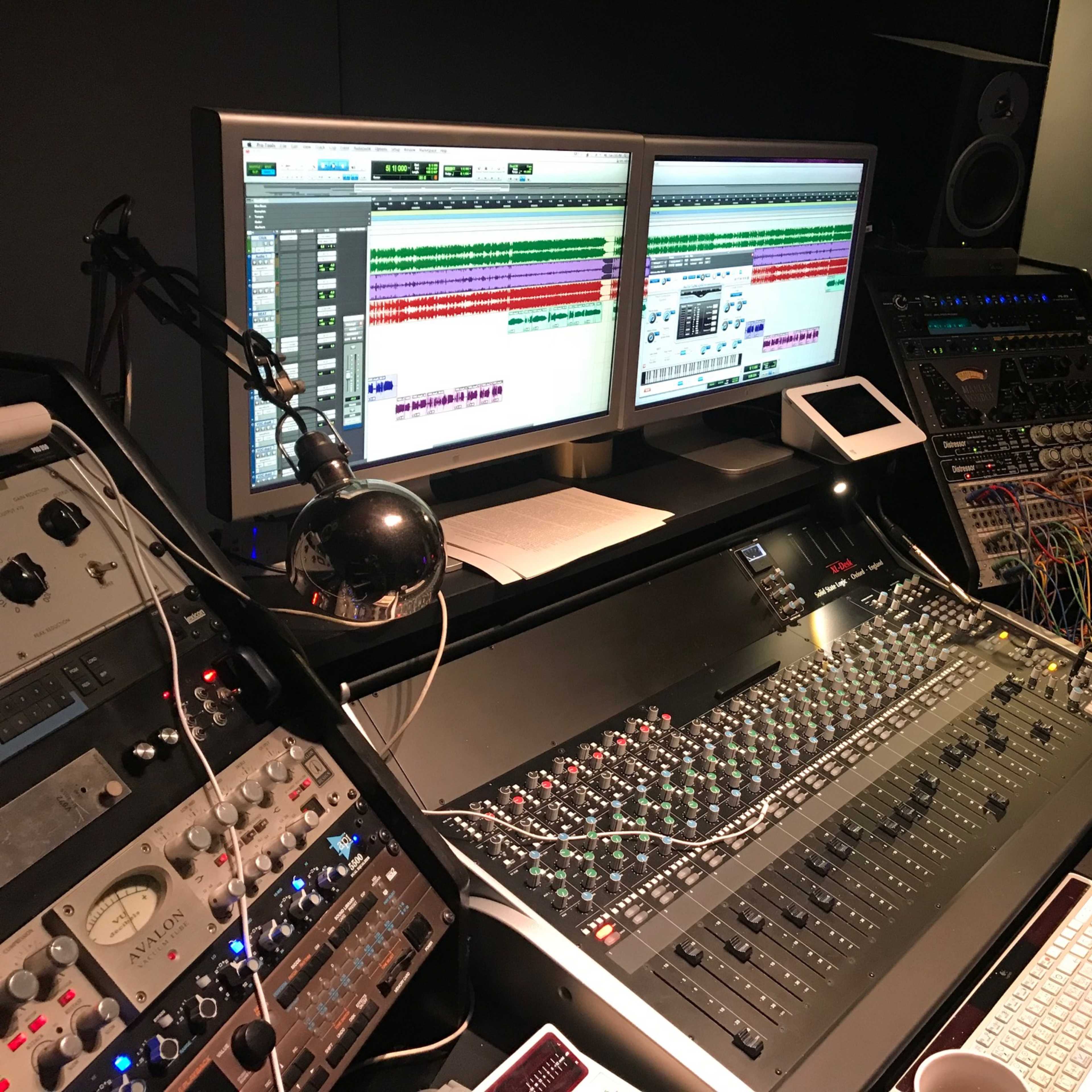 A music studio with a mixing console, two computer monitors displaying a digital audio workstation, and various audio equipment and cables.