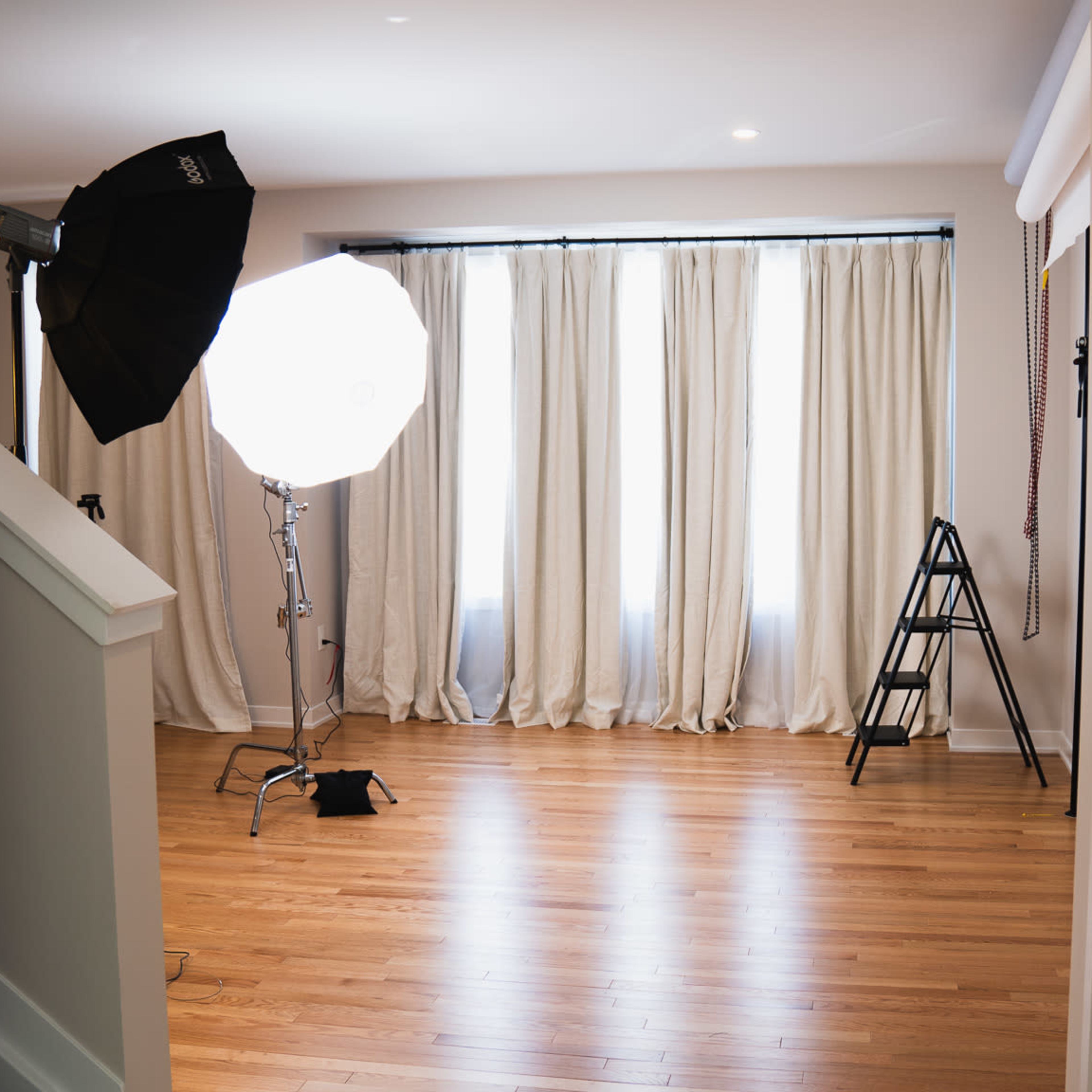 A bright, open room with wooden flooring, large windows covered by white curtains, and photography equipment set up in one corner.