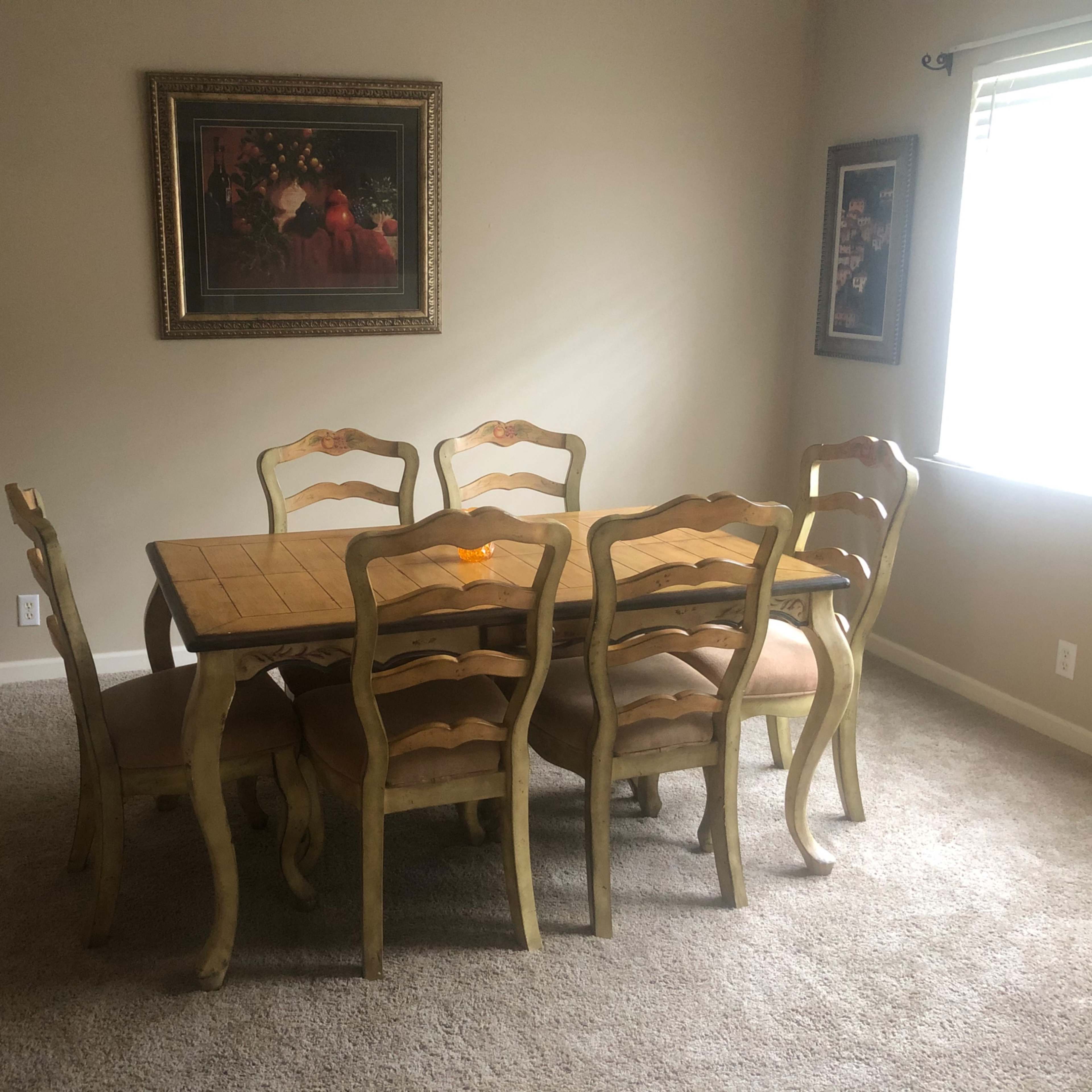 A wooden dining table surrounded by six chairs is positioned in a carpeted room with two framed pictures on the walls and a window that lets in natural light.